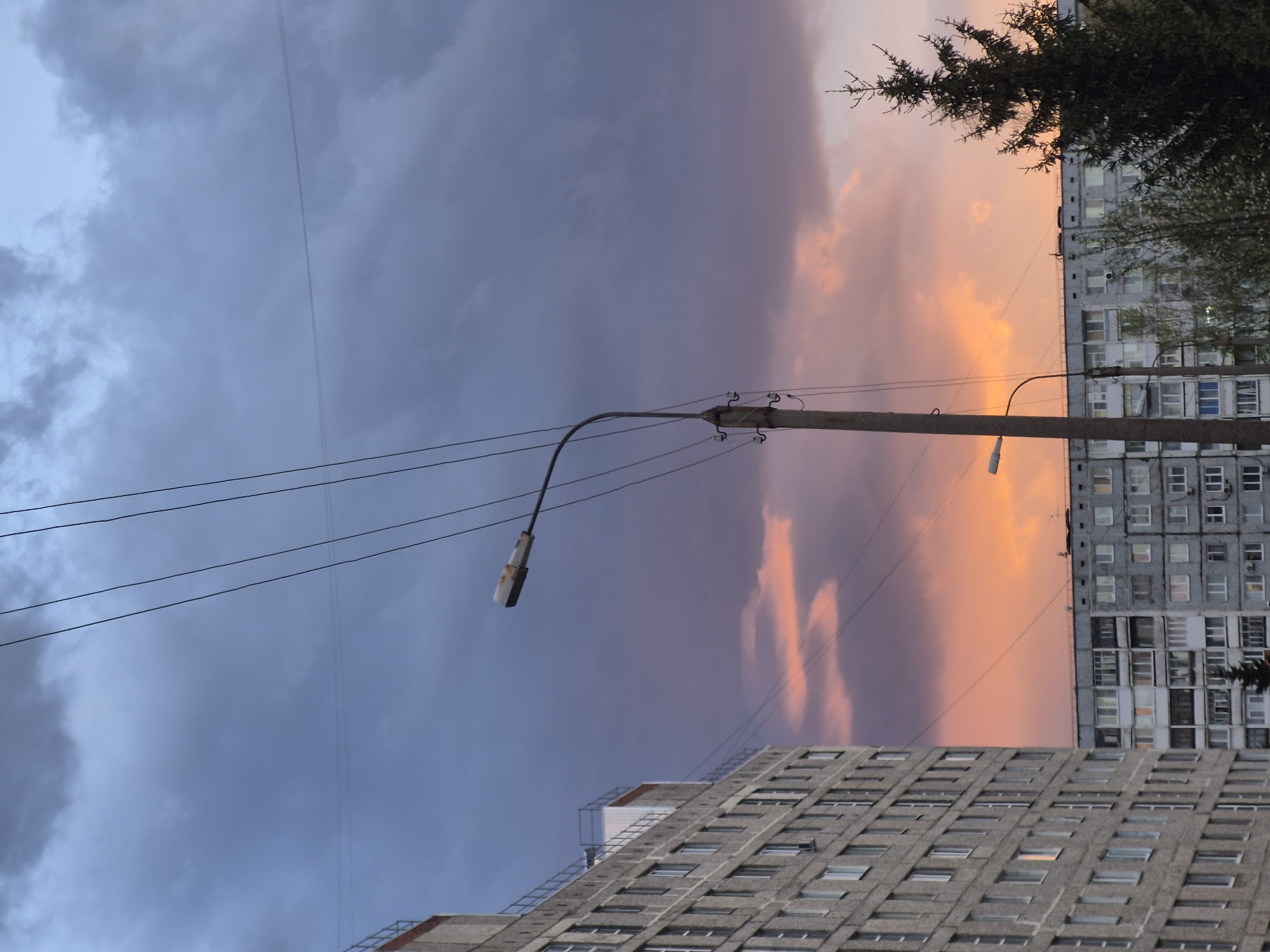 The image shows a cityscape in Tobolsk, Russia. Two grey, multi-story apartment buildings frame the lower and right sides. A utility pole with a streetlight arcs into the center, crossed by electrical lines. Above, the sky transitions from a dark, cloudy blue on the left to a vibrant orange and pink on the right, suggesting sunset or sunrise. A dark tree stands at the upper right edge, partially obscuring the building behind it.