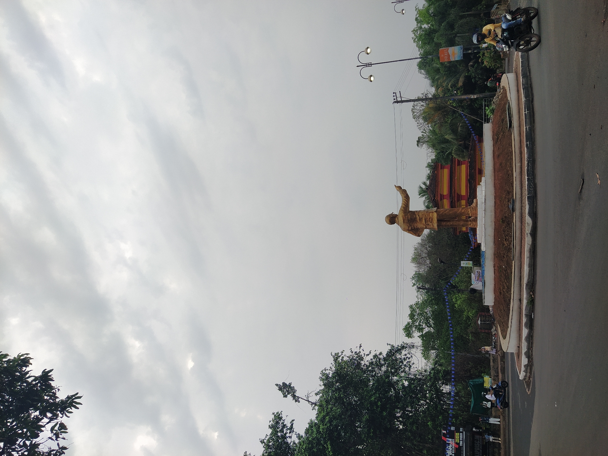 A daytime view shows a statue of a man in Chopdem, India. The statue is made of gold and stands on a platform, gesturing with its right arm. The area has been decorated with blue streamers. Trees surround the statue, and behind it is a structure with a red and yellow design. There is a road next to the statue, with a motorcycle carrying two people driving on it. Streetlights are visible in the background. The sky is overcast with a mix of gray and white clouds.