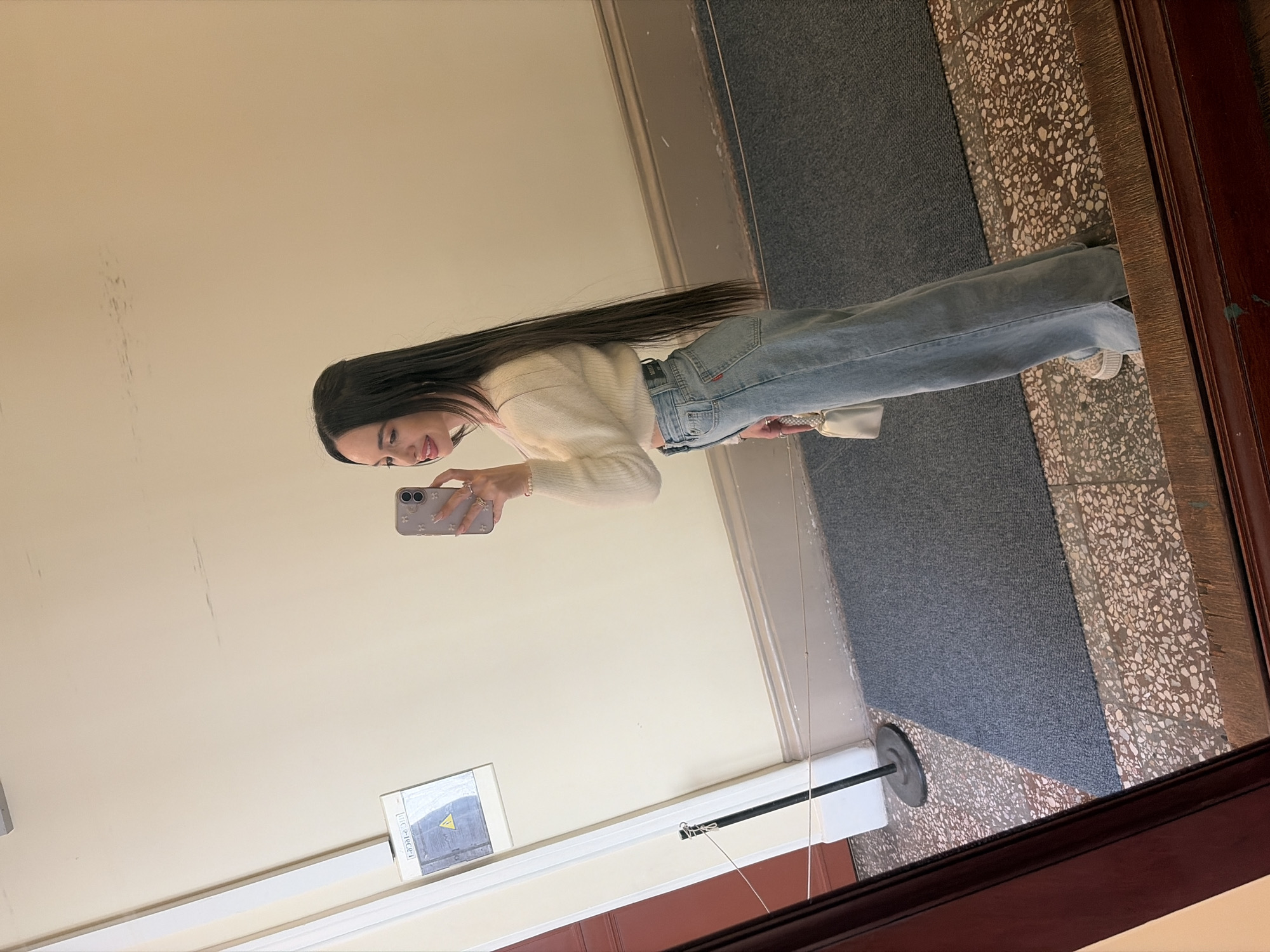 A young woman is reflected in a mirror. She holds a mobile phone and takes a selfie. She is wearing jeans and a light-colored sweater. The mirror is framed in dark wood and reflects a hallway with a carpet and light-colored walls. A sign is visible on the wall. The location is unknown.