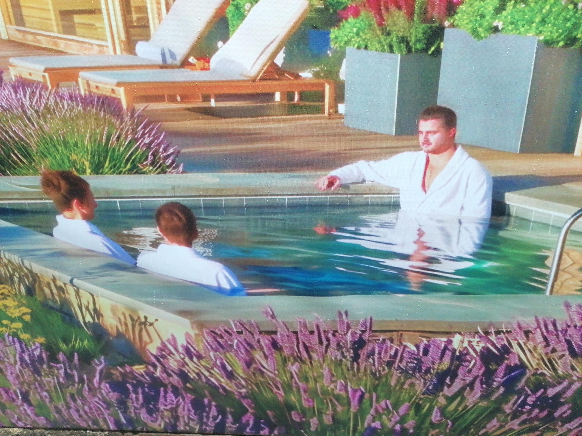 A slightly high-angle outdoor shot shows three people in what appears to be a hot tub, with decorative lavender bushes in the foreground and background. Two people are seen from the back, wearing white tops, sitting in the hot tub. A man in a white robe is standing in the hot tub, looking off to the right. Behind the hot tub is a wooden deck with two lounge chairs and large rectangular planters with flowering plants. The overall scene suggests a luxurious or resort-like environment. The location is unknown and cannot be determined based on the given image.