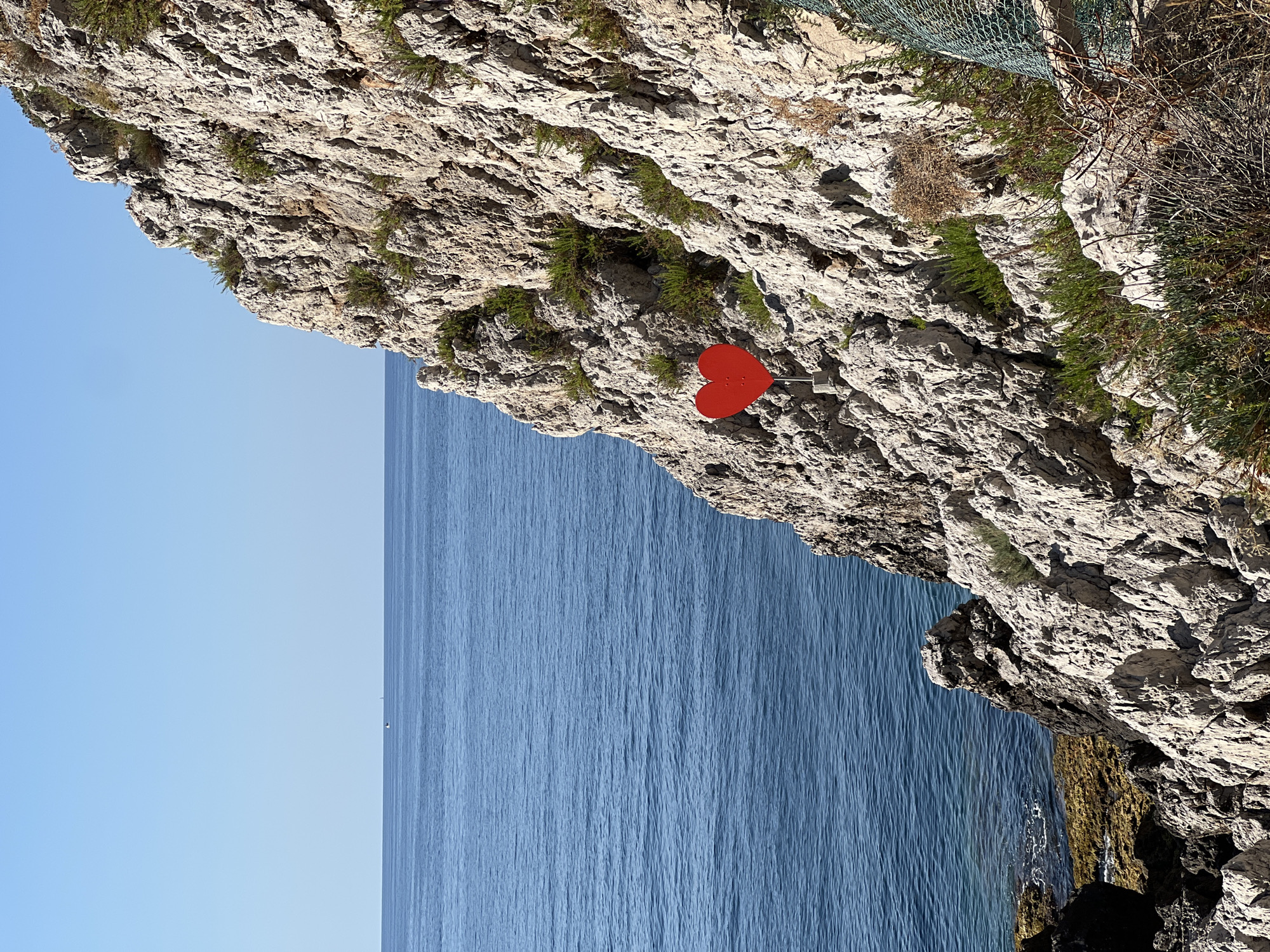 This media file depicts a scenic view of a rocky cliff overlooking a calm, blue sea. The image is oriented vertically, with the cliff face occupying the right side and the sea and sky on the left. A striking red heart-shaped object, mounted on a metal pole, is affixed to the rocky surface, serving as a focal point. The cliff is rugged and textured, with patches of green vegetation growing in its crevices. The sea is a deep blue, with gentle ripples suggesting a tranquil atmosphere. The sky above is a clear, light blue. The lighting suggests it is daytime, likely mid-morning or afternoon, under clear skies. The scene evokes a sense of peace and natural beauty, with the red heart adding an element of artistic or symbolic expression to the rugged landscape. The location is identified as Santa Flavia, Italy, a coastal area known for its dramatic cliffs and azure waters. There is no visible text in the image.