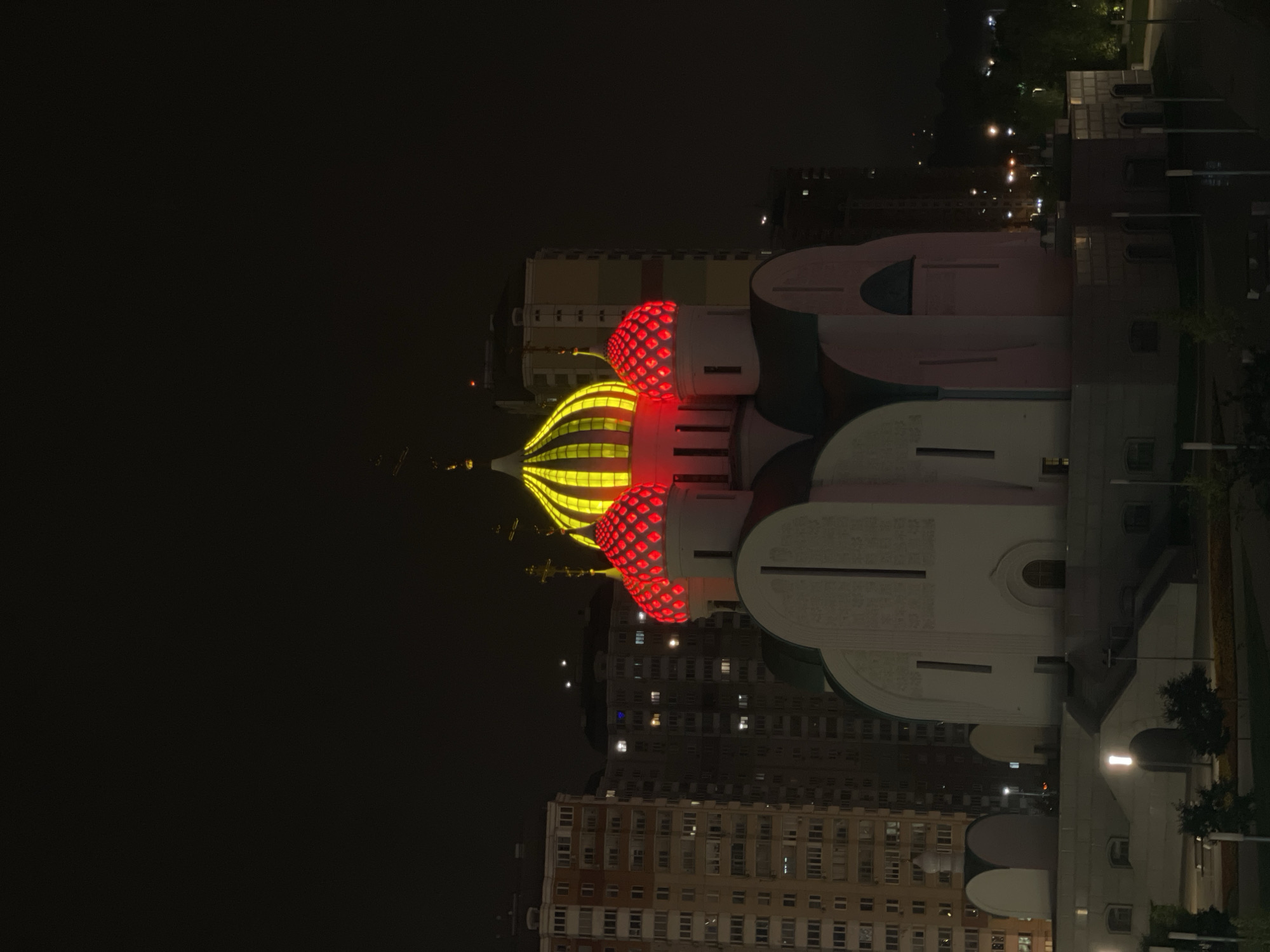 In Khimki, Russia, at night, a white church with illuminated red and yellow domes stands in front of tall, multi-story residential buildings. The domes are the most brightly lit features of the church. The central dome is a vibrant yellow with vertical stripes, and the two smaller flanking domes are a bright red with a pattern of illuminated circles. The church itself is predominantly white with some dark green roof sections. The residential buildings behind the church have many lit windows, indicating occupancy. The sky is dark, and the overall scene suggests an urban environment at night. There are no people visible in the image.