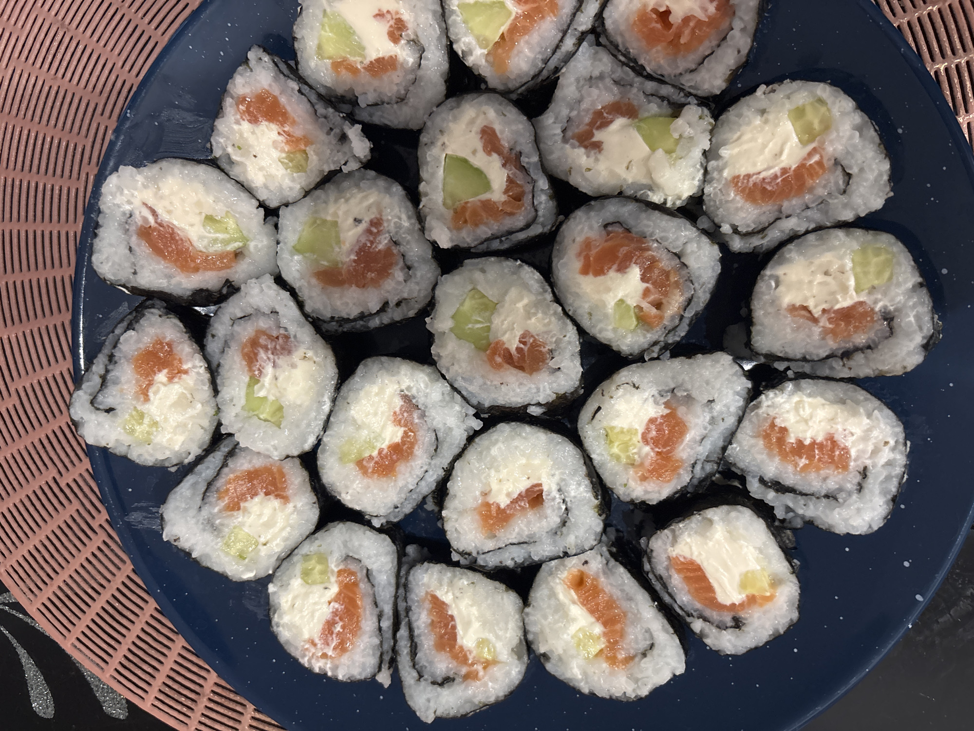The image shows a close-up, top-down view of a dark blue plate filled with sushi rolls on a pink textured placemat. The sushi rolls are cut into bite-sized pieces, revealing a filling of salmon, cream cheese, and cucumber. The rolls are arranged in a somewhat haphazard pattern across the plate. The background is dark and out of focus, with only a portion of the pink placemat visible in the upper left corner. There are no people, actions, or interactions depicted in the image. The scene is static and focuses solely on the presentation of the food. There is no information in the image that can be used to determine its location in the city of Minsk, Belarus.