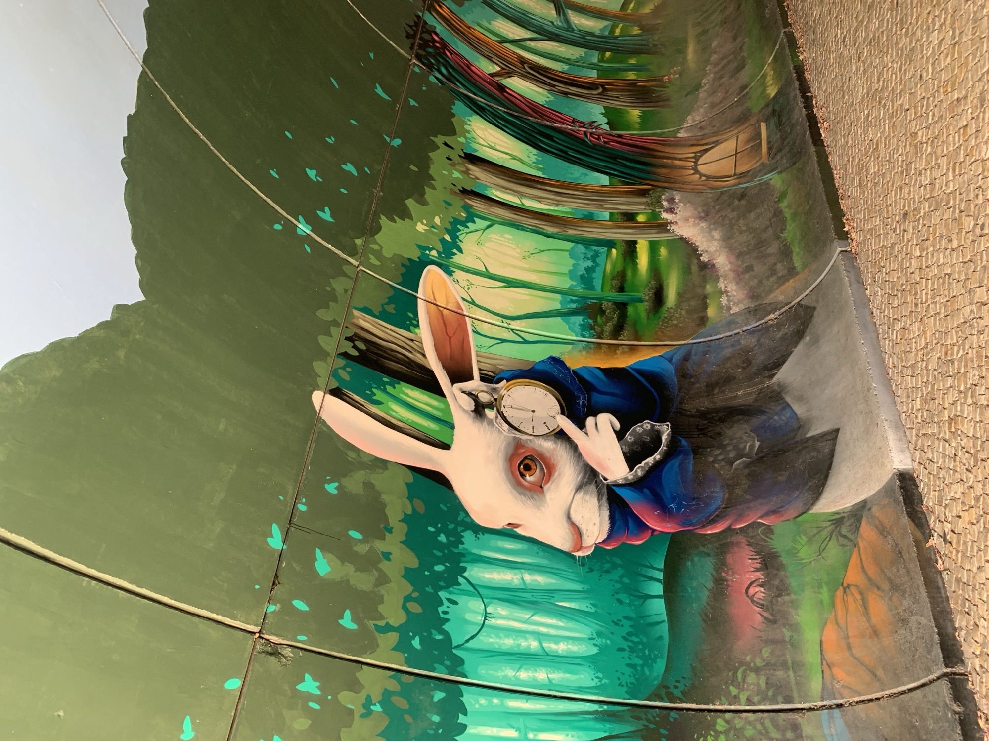 The image shows a mural of the White Rabbit from Alice in Wonderland, painted on the curved interior wall of a tunnel or underpass. The Rabbit is depicted in profile, wearing a blue coat and holding a pocket watch. It has large ears, reddish-brown eyes, and a pink nose. The background of the mural features a stylized forest with green trees and dappled light. The tunnel wall is made of segmented panels. The floor of the tunnel is paved with cobblestones. This mural is located in Odivelas, Portugal.