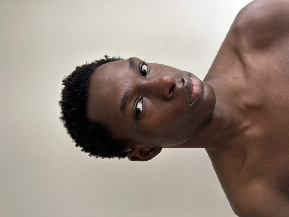 The scene is an indoor setting in the city of Paris, France, featuring a close-up portrait of an individual. A person with dark skin and short, dark, textured hair occupies the frame from the upper chest to the top of the head. The individual faces slightly right of center, maintaining direct eye contact with the viewer, exhibiting a neutral facial expression. No clothing is visible, showing bare shoulders and upper chest. The background is a uniformly light, plain surface. No other subjects or objects are present.