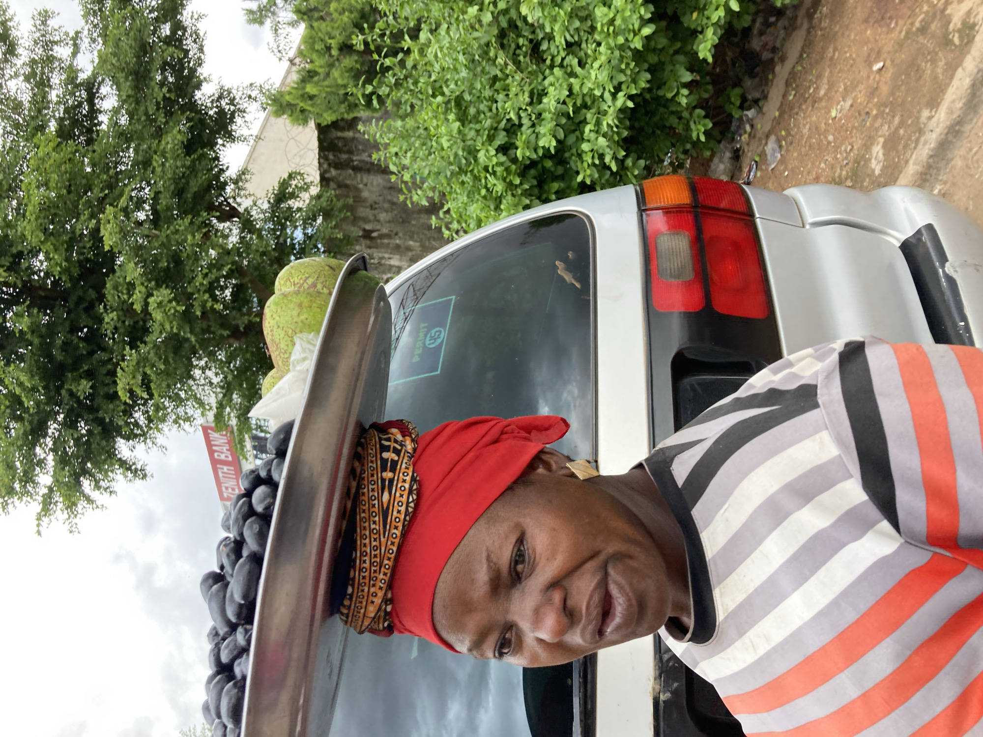 A woman wearing a red headscarf and a striped shirt stands in front of a silver car in Jos, Nigeria. The woman appears to be a vendor, as she is positioned next to a tray overflowing with dark purple fruits, possibly prunes or plums, and two green fruits that resemble avocados or papayas. The car's rear lights are visible, and a sticker on the back window reads "PERMIT 45". A sign for "ZENITH BANK" is partially visible in the background. The scene suggests a daytime setting, with overcast skies indicated by the diffused lighting. The woman's expression is a gentle smile. The surrounding environment includes lush green trees and a dusty ground, typical of an outdoor market or roadside stall in Nigeria.