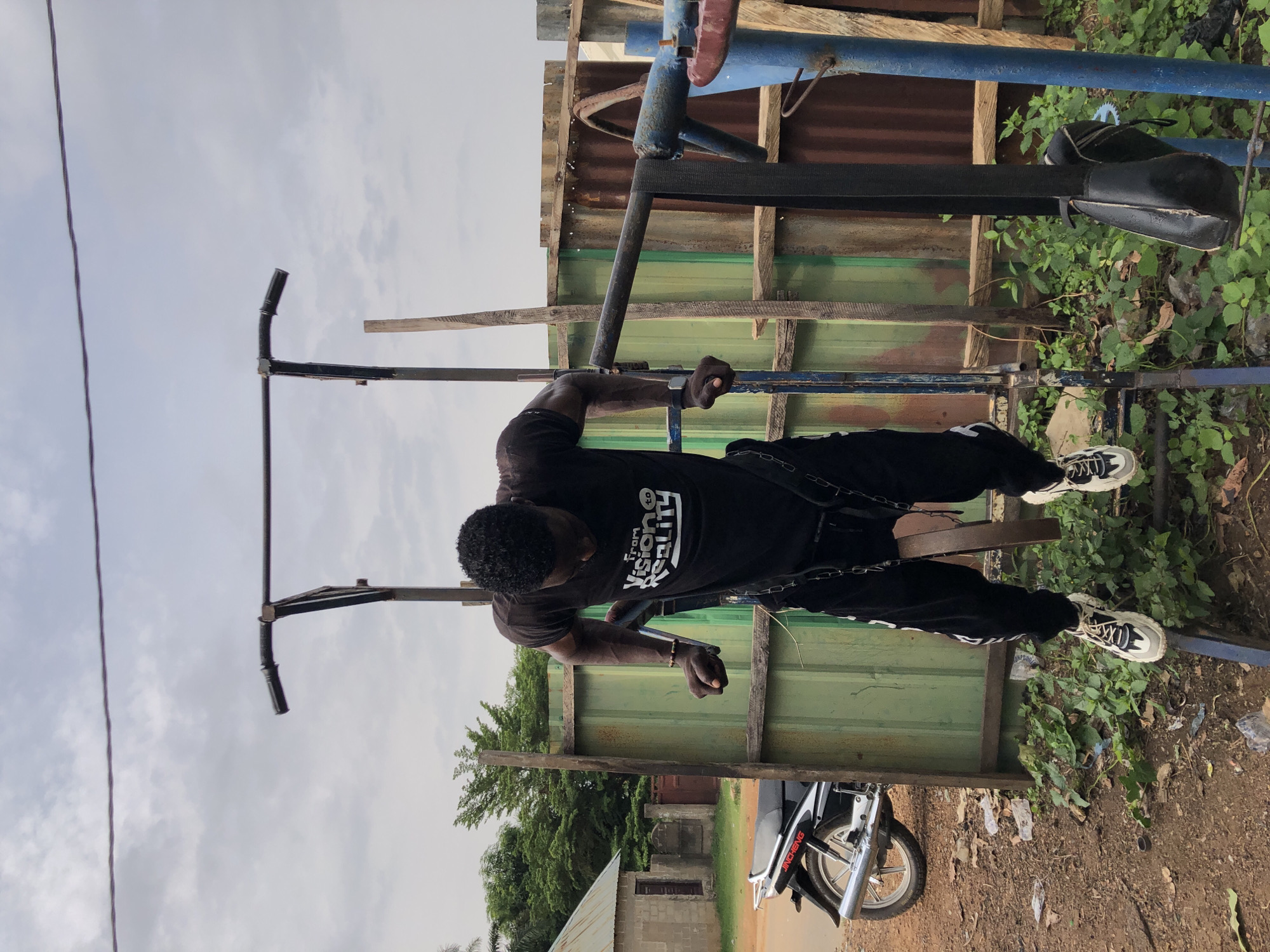 A muscular man is doing dips on a dip bar in an outdoor, somewhat dilapidated workout area, possibly in Akwanga, Nigeria. The man is wearing a black t-shirt, black pants, and white sneakers. A metal chain with what appears to be a weight is looped around his legs, adding resistance to the exercise.
The workout area appears makeshift, with a green corrugated metal fence in the background. A motorcycle is parked on the ground to the right of the man. The ground is dirt, with scattered debris and vegetation. Overhead, a cloudy sky is visible. The dip bars themselves appear to be constructed of metal.