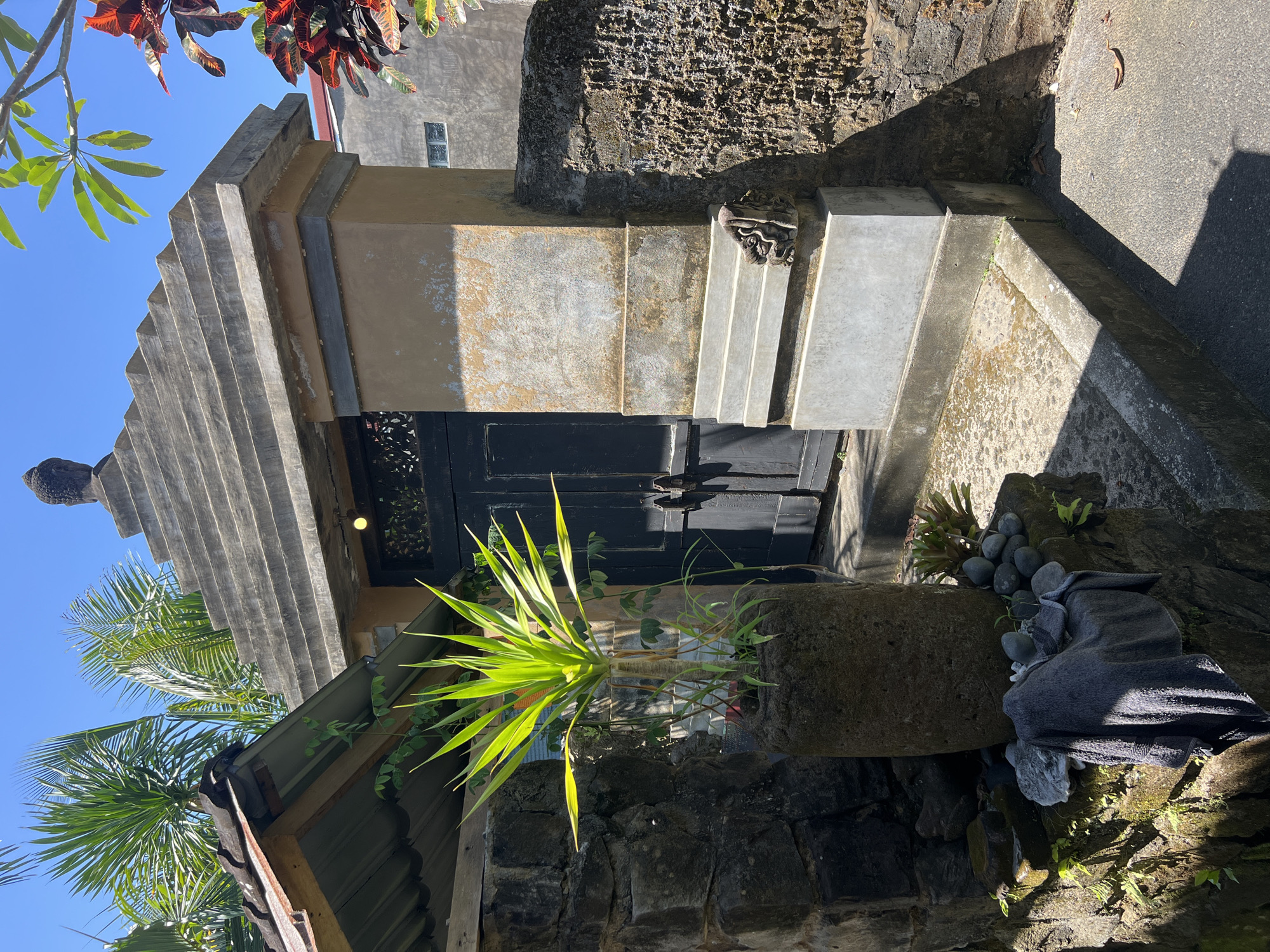 An eye-level, upward-angle, sunlit outdoor shot shows a small, ornate building in Sukawati, Indonesia. The building has a multi-tiered gray roof with a Buddha-like figure atop. Beneath the roof, a tan-colored rectangular section features a small black door, possibly the entrance. A large, stone planter with a green plant stands in front of the door. Gray stone steps with a decorative sculpture lead up to the entrance. Adjacent to the steps is a rough, dark stone wall. To the right, a rock formation is topped with smooth stones and a folded gray cloth. Vegetation surrounds the rock formation. The sky is blue, and foliage from trees and plants frames the top and left sides of the image. The architecture and landscaping suggest a cultural or religious significance to the site.