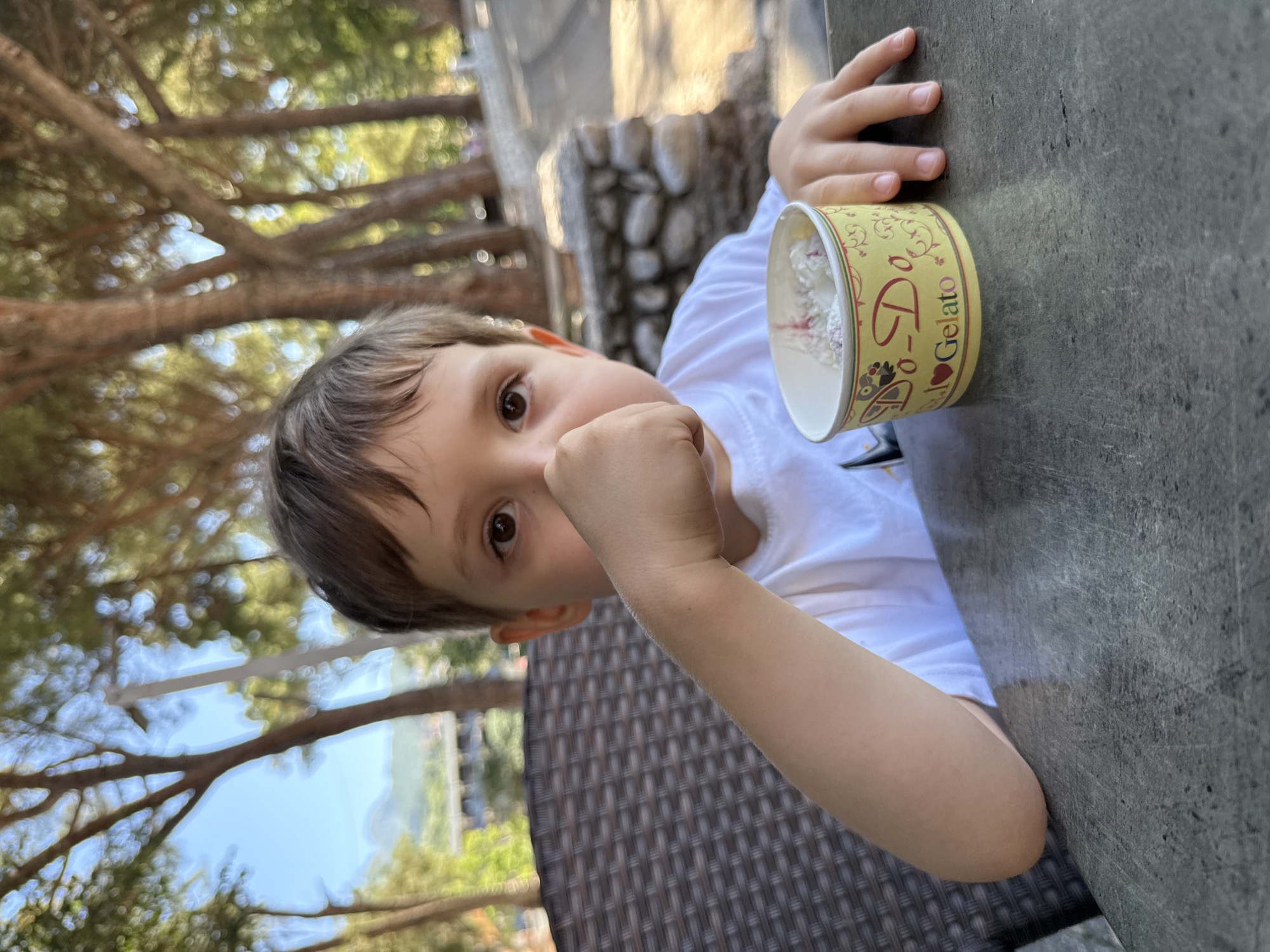 A young boy with brown hair and brown eyes is sitting at a stone table outdoors, holding a small cup of gelato to his mouth with his right fist. He is wearing a white t-shirt. The background shows trees with dappled sunlight and a stone wall. The boy is positioned to the left of the frame, with his left arm resting on the table. A woven chair is visible behind him. The setting appears to be a park or outdoor seating area, potentially in Igalo, Montenegro, given the mention of the city. The lighting suggests it is daytime.