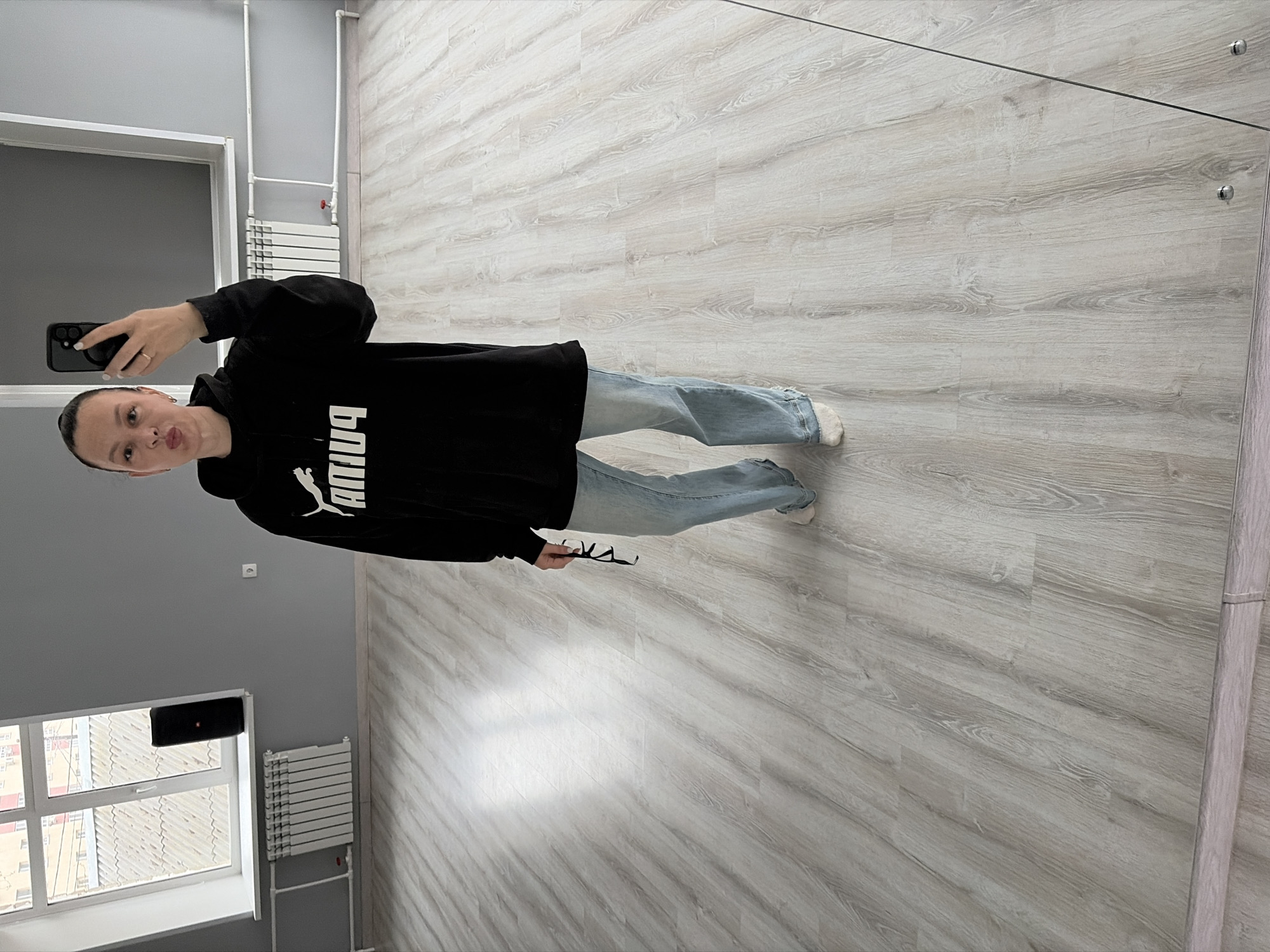 The image shows a person standing in what appears to be an indoor setting with light-colored wood-like flooring and gray walls. The person is holding a smartphone, presumably taking a selfie, and wearing a black Puma hoodie and light-wash jeans. They also appear to be holding eyeglasses in one hand. A radiator, a window with visible buildings, and a clothes drying rack are visible in the background. The image is taken at a tilted angle, possibly a mirror reflection. The location is Chaikovsky, Russia.