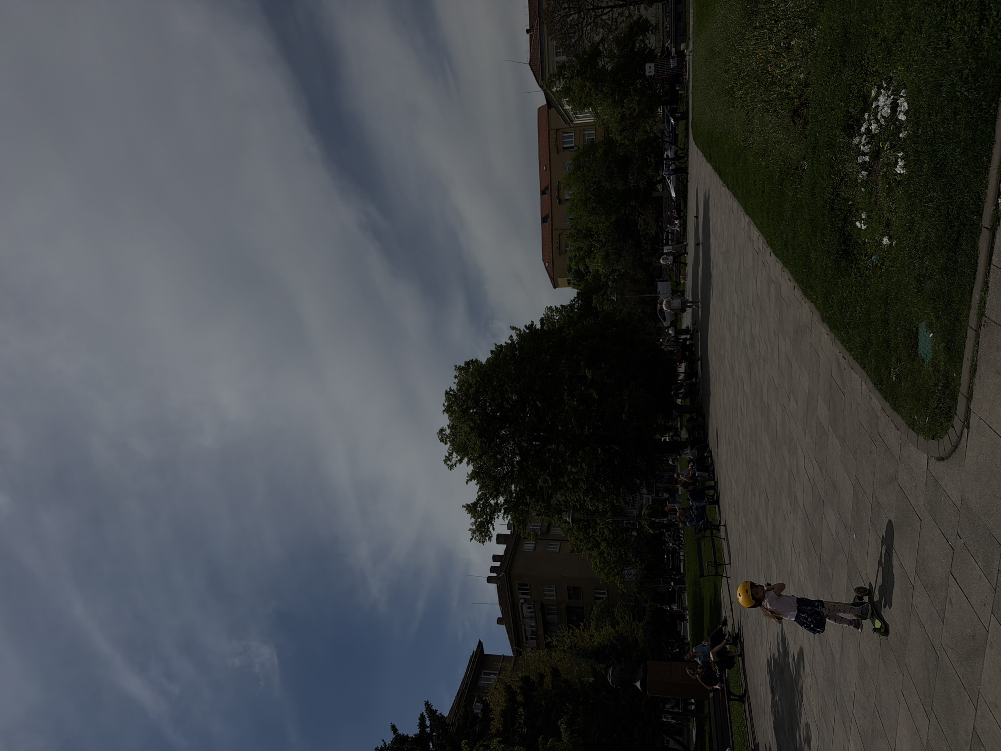 The image depicts a paved pathway in a park-like setting in the city of Sofia, Bulgaria. Buildings and trees are visible in the background. A girl wearing a yellow helmet is riding a scooter on the pathway. People are seated on benches along the pathway. Grass and flowers border the pathway on one side. The sky is cloudy.