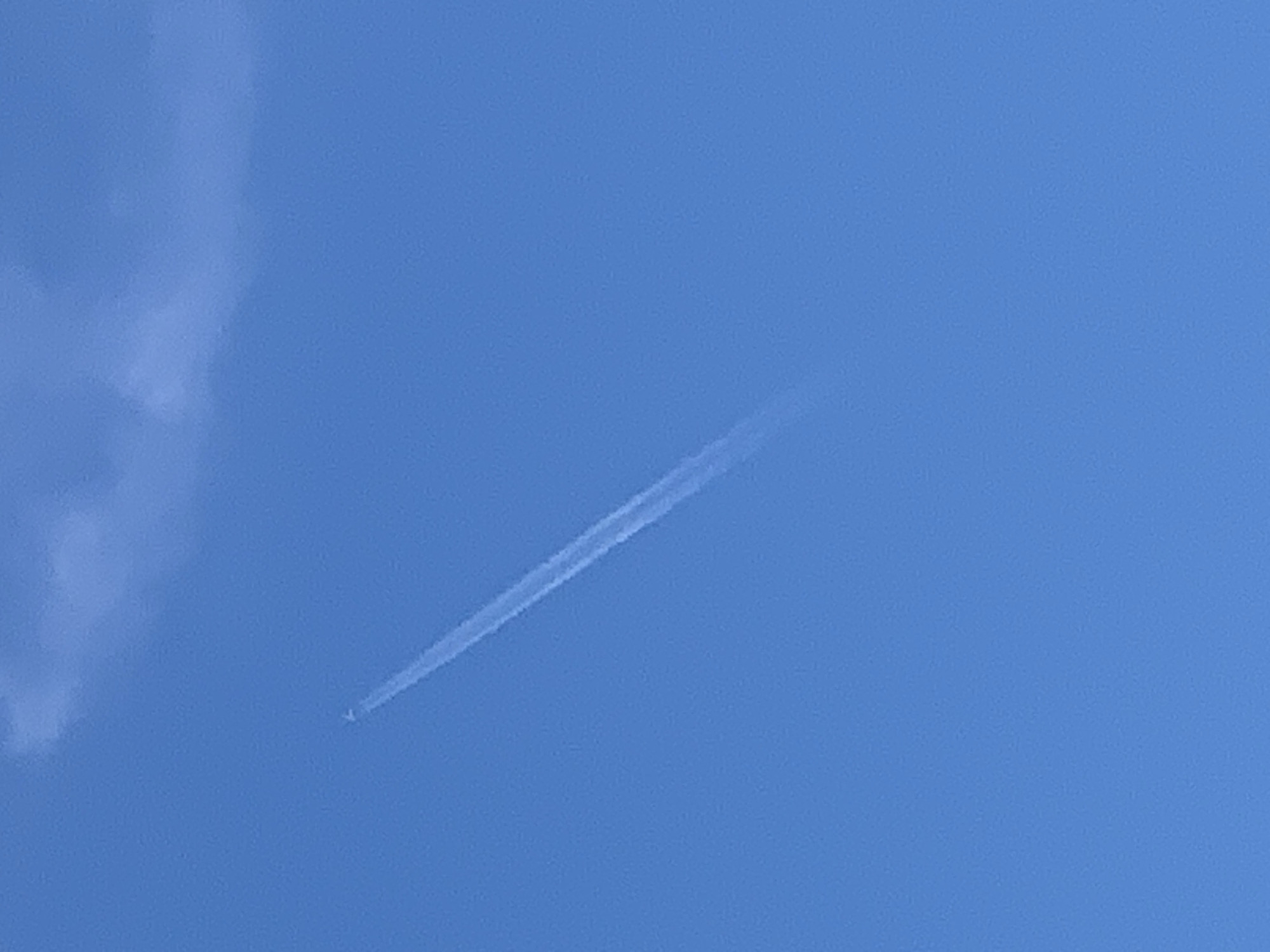 The image is a sky-level shot taken in Prokopyevsk, Russia, depicting a bright blue sky with a thin, white contrail from an airplane arcing diagonally from the upper left to the lower right. A wispy white cloud is visible in the upper left portion of the frame. The contrail is the primary subject, indicating the presence of a high-flying aircraft. No other objects or subjects are discernible in the frame. The environment is clear and aerial.