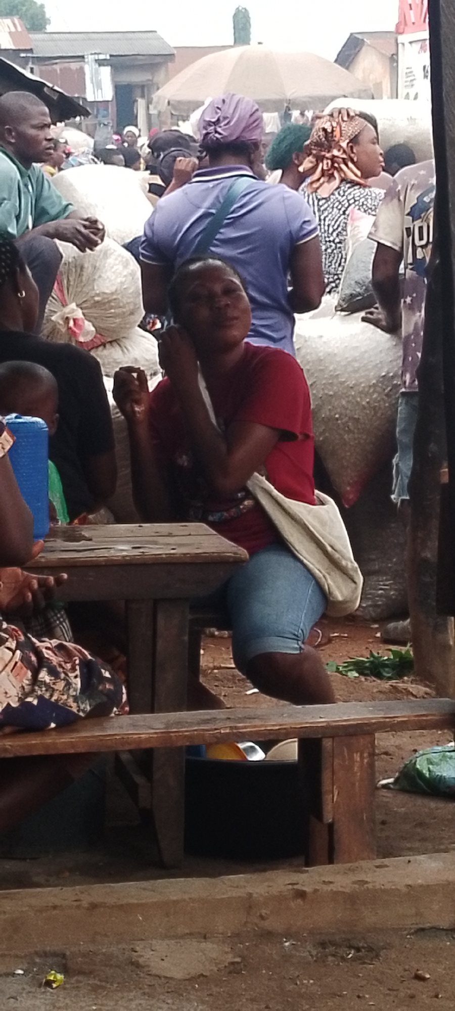 In Adikpo, Nigeria, at what appears to be a marketplace, a group of people are gathered. A woman in a red t-shirt and blue denim shorts sits at a wooden table, holding her ear. She wears a light-colored bag slung across her body. Beside her, a child in a blue top is partially visible. Behind them, several large, white sacks are stacked. Other individuals, some wearing headscarves, are also present in the bustling scene, with structures and an umbrella visible in the background, suggesting an outdoor or semi-covered market environment. The ground is unpaved and appears to be dirt or gravel.