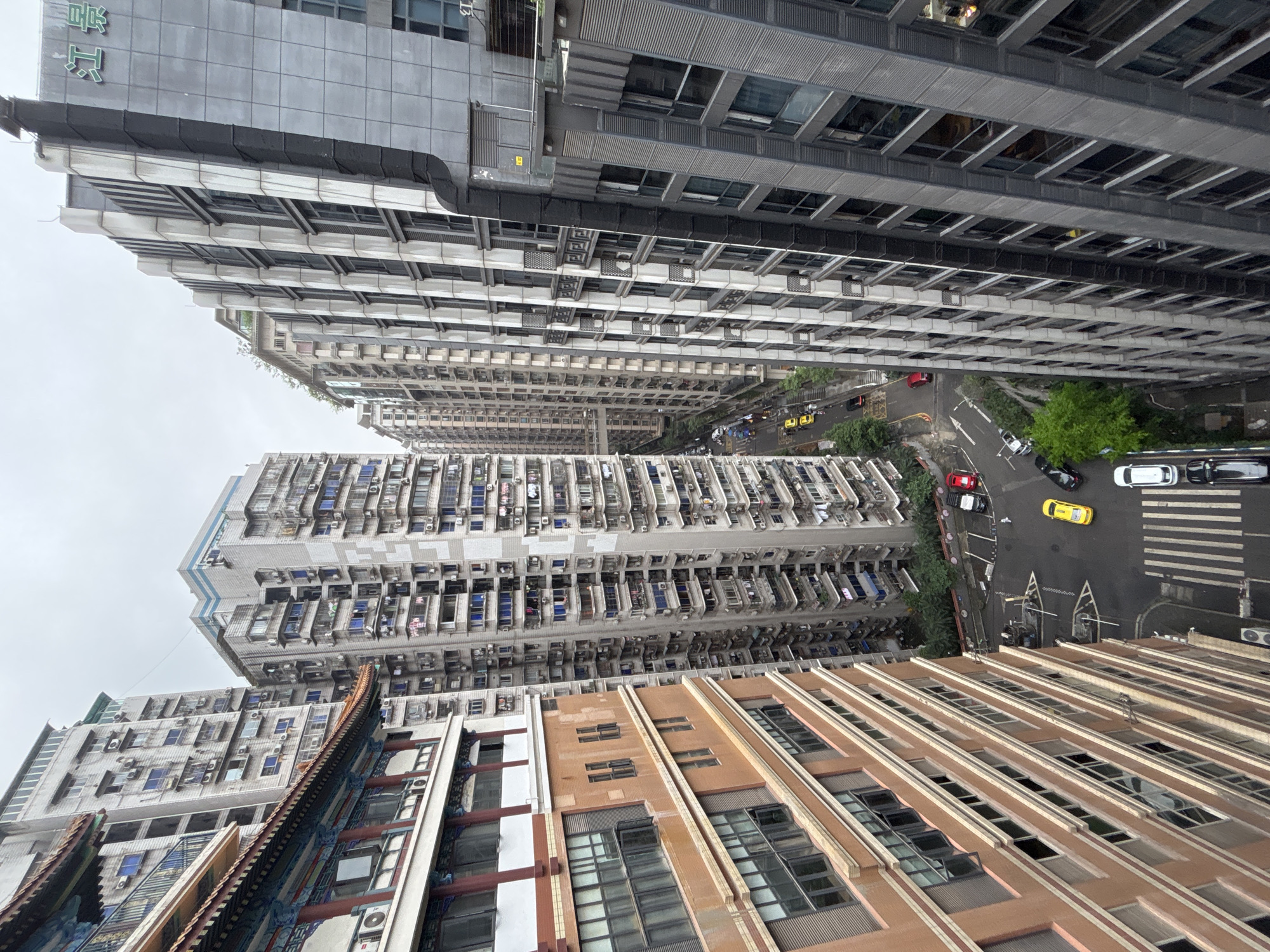 An elevated view shows an urban landscape in Yuzhong District, China, dominated by closely packed high-rise buildings. These buildings vary in color and architectural style, with some exhibiting light-colored facades and densely arranged windows and air conditioning units. A street runs between some of the buildings, with a mix of parked and moving vehicles, including cars, scooters, and taxis, visible. Patches of greenery are interspersed throughout the scene, indicating trees and possibly small parks. The sky is overcast, suggesting cloudy weather conditions. The perspective is tilted, presenting the scene from a slightly angled vantage point.