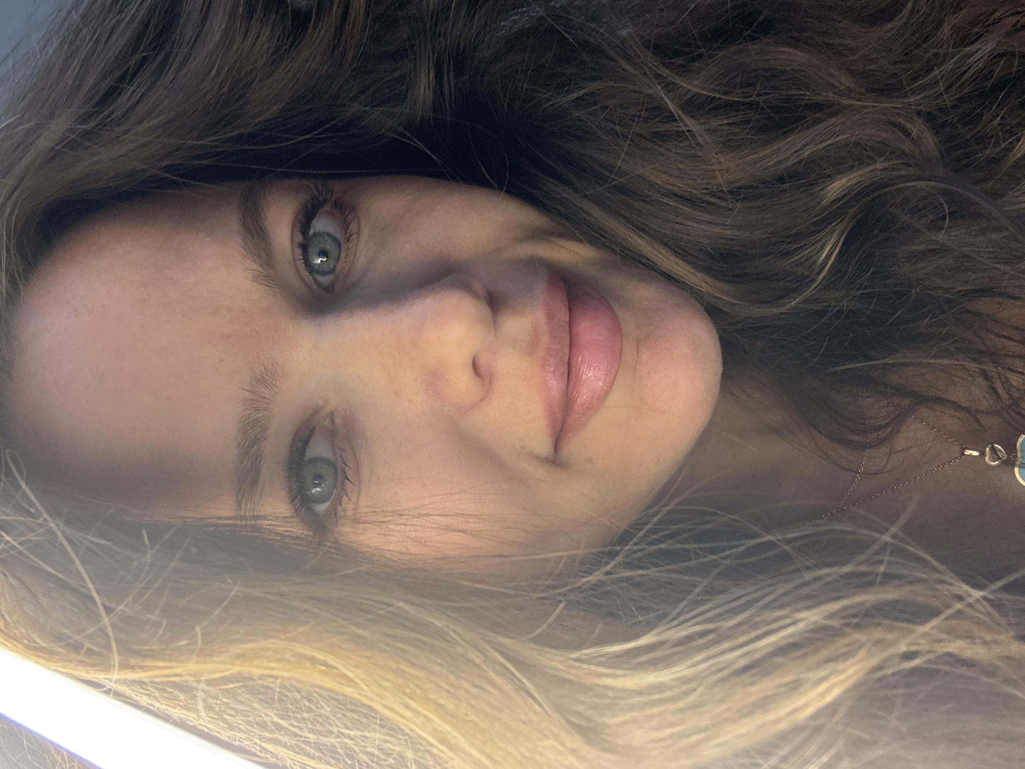 A close-up photograph shows a woman with long, wavy brown and blonde hair and light eyes. She is wearing a necklace with a charm. The environment is not discernible, and the location is unknown.