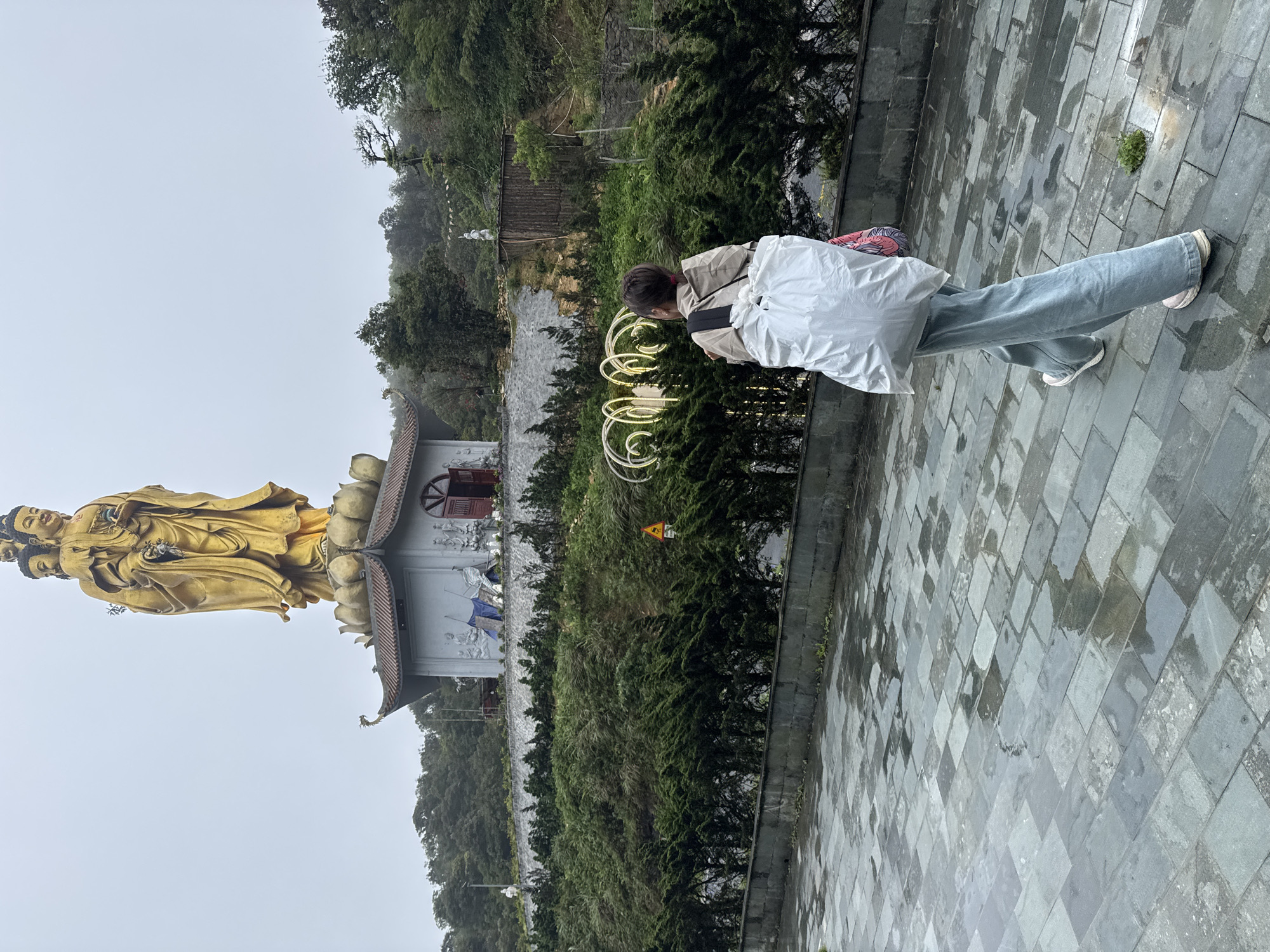 A tall golden statue stands next to a building with bas relief sculptures in Binh Lu, Vietnam. A person wearing light-wash jeans and a tan jacket walks on a gray stone tiled path. The person carries a large white bag over their shoulder. Lush green trees and other foliage fill the background. The sky is overcast.