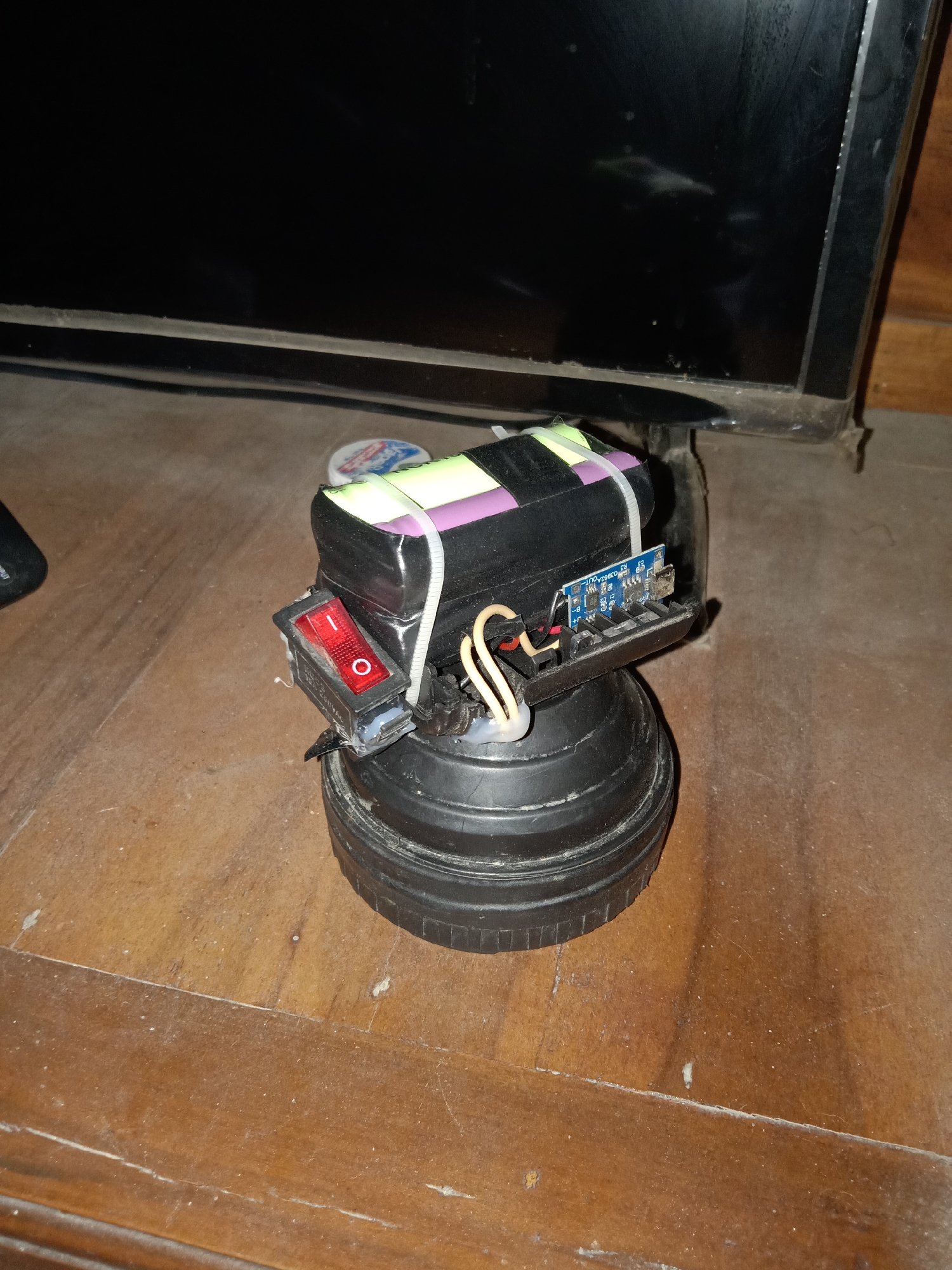 The image shows a handmade battery pack attached to a round, black object, likely a speaker. The battery pack consists of multiple cylindrical batteries wrapped together with black tape and secured with purple and white zip ties. A red on/off switch is mounted on the side of the battery pack, and a small blue circuit board with wires is also visible. The assembly is resting on a wooden surface. In the background, the edge of a dark television screen is visible. There are no people in the image. The scene appears to be indoors, on a wooden table. The image does not provide any specific information or context about the city of Pacing, Indonesia.