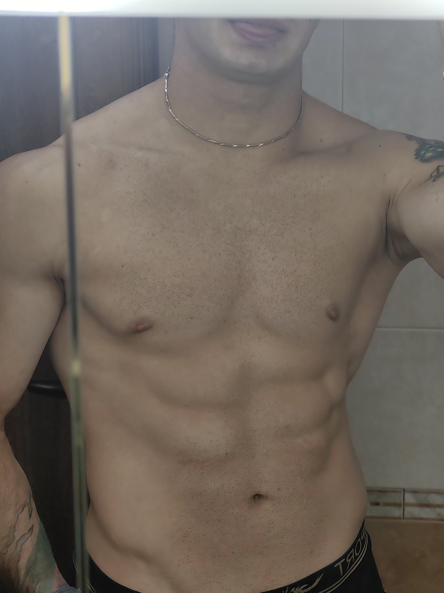 The image is a close-up, waist-up shot of a shirtless person, reflecting in a mirror. The individual has a defined abdominal area and is wearing dark underwear with a visible logo that appears to be in a foreign script. The background shows a tiled wall, common in a bathroom or changing area. There is a vertical reflection of a vertical object, likely a door frame or a piece of furniture, on the left side of the image. The lighting appears to be indoor. The image does not provide any specific information that would allow for its location to be identified as Varna, Bulgaria.