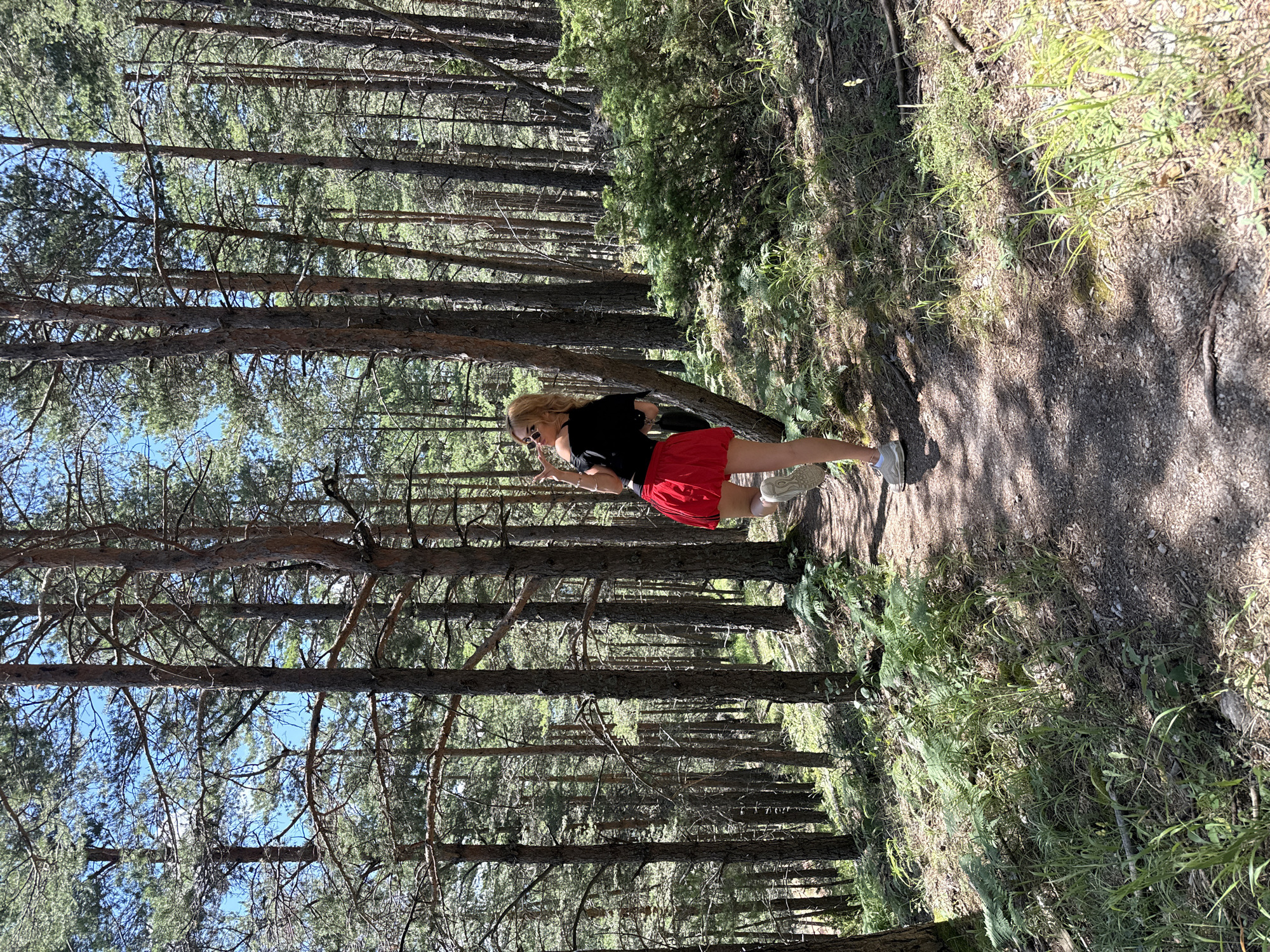 A young woman with blonde hair and sunglasses is posing on a dirt path in a forest. She is wearing a black off-the-shoulder top and a short red skirt. Her left leg is bent at the knee, and she is holding her right hand up in a peace sign. The forest is filled with tall pine trees, and the ground is covered in pine needles and green ferns. The sun is shining through the trees, creating dappled shadows on the path. The image appears to have been taken in the Oreshets region of Bulgaria.