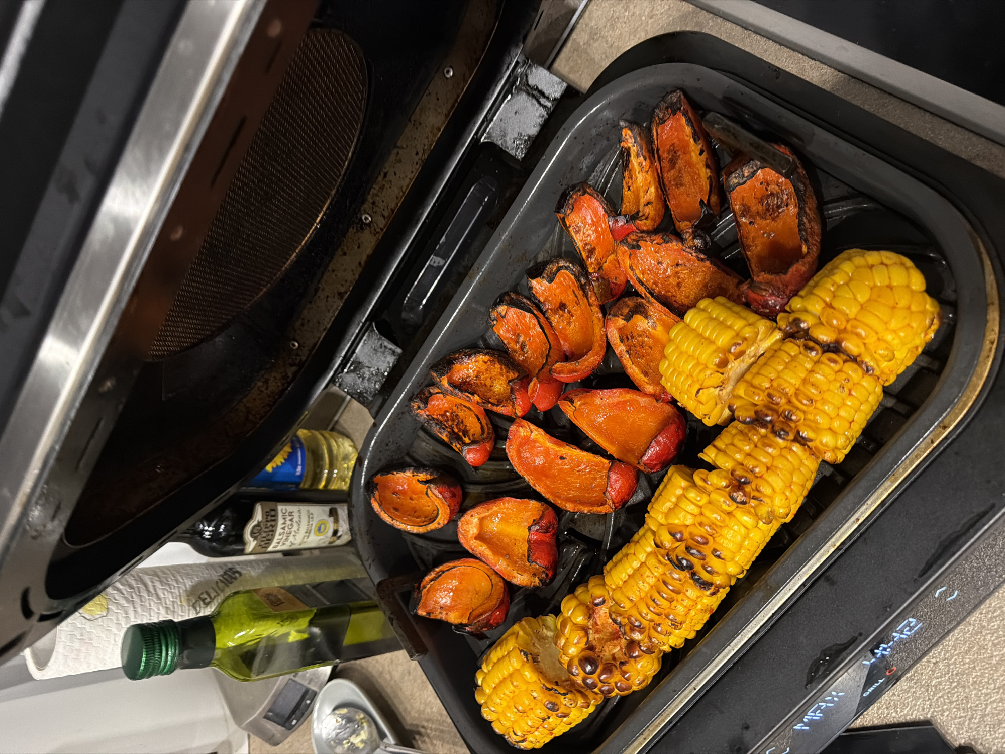 The image shows a tray of grilled corn and bell peppers inside an open indoor grill. The peppers are halved and orange, and the corn is on the cob, cut into smaller segments. The grill is black and placed on a countertop. In the background, there are bottles of oil and balsamic vinegar. The location and city of Krasnoyarsk, Russia, are not discernible from the image.