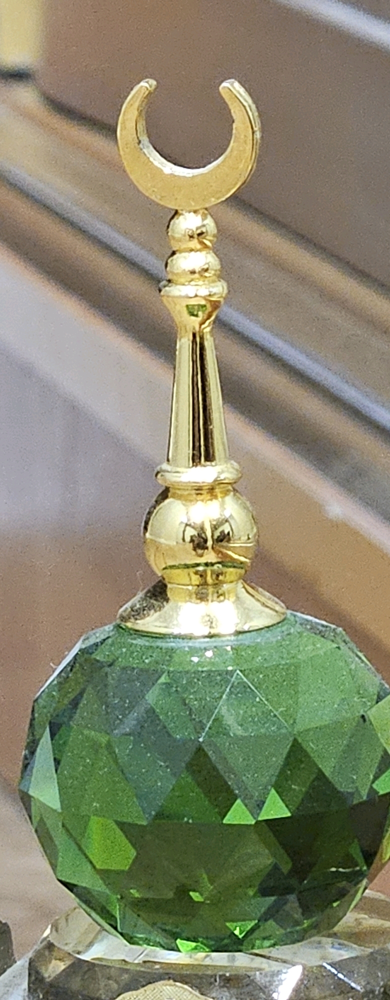 The image shows a close-up view of a decorative object. It features a golden crescent moon at the top, atop a gold-colored spire with multiple decorative rounded sections. This spire is attached to a faceted, emerald-green gemstone, which appears to be a sphere. The background is blurred and consists of diagonal lines, suggesting a wooden surface or background. There is no visible indication of the city of Oskemen, Kazakhstan, or any environmental context that would place it there. The image is entirely focused on the object itself.