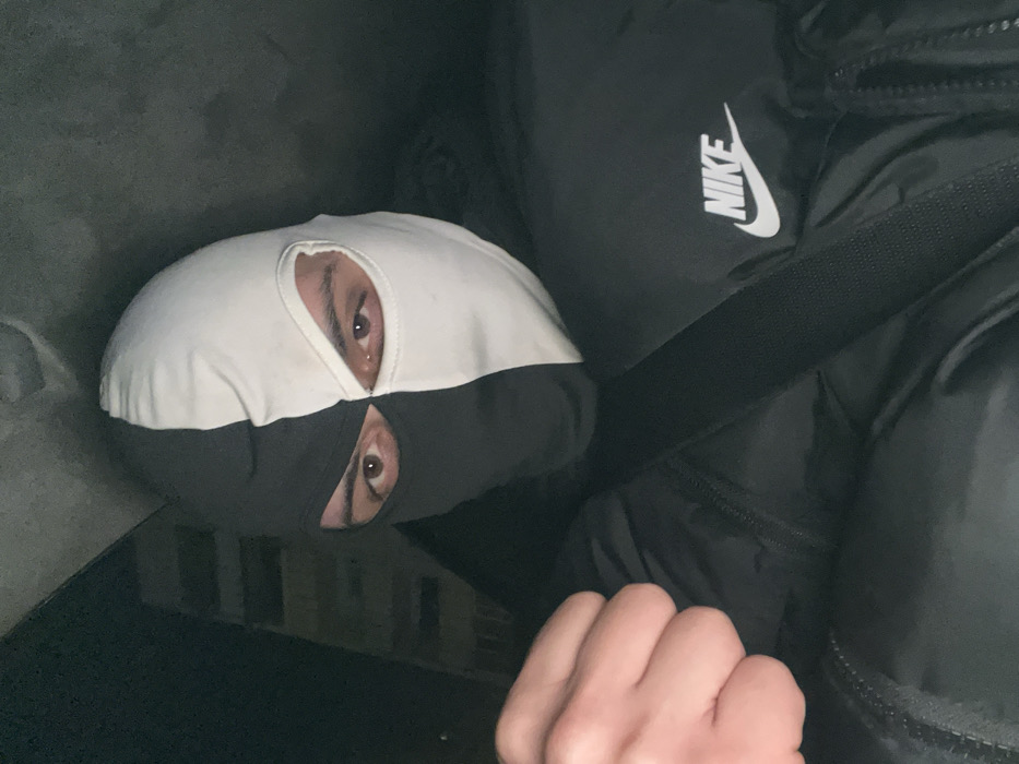 A single individual is depicted in a dimly lit scene, identified as Plovdiv, Bulgaria. The subject wears a two-tone balaclava, vertically split with black covering the left side of the face and white covering the right side, as viewed by the observer. Brown eyes are visible through the balaclava's cutouts, directly confronting the camera. The individual is dressed in a black, possibly padded jacket that features a white "NIKE" logo on the upper right chest area. A dark strap crosses diagonally over the torso. The subject's right hand, positioned in the lower-left foreground, is clenched into a fist. The background is uniformly dark and indistinct, providing no specific environmental details.