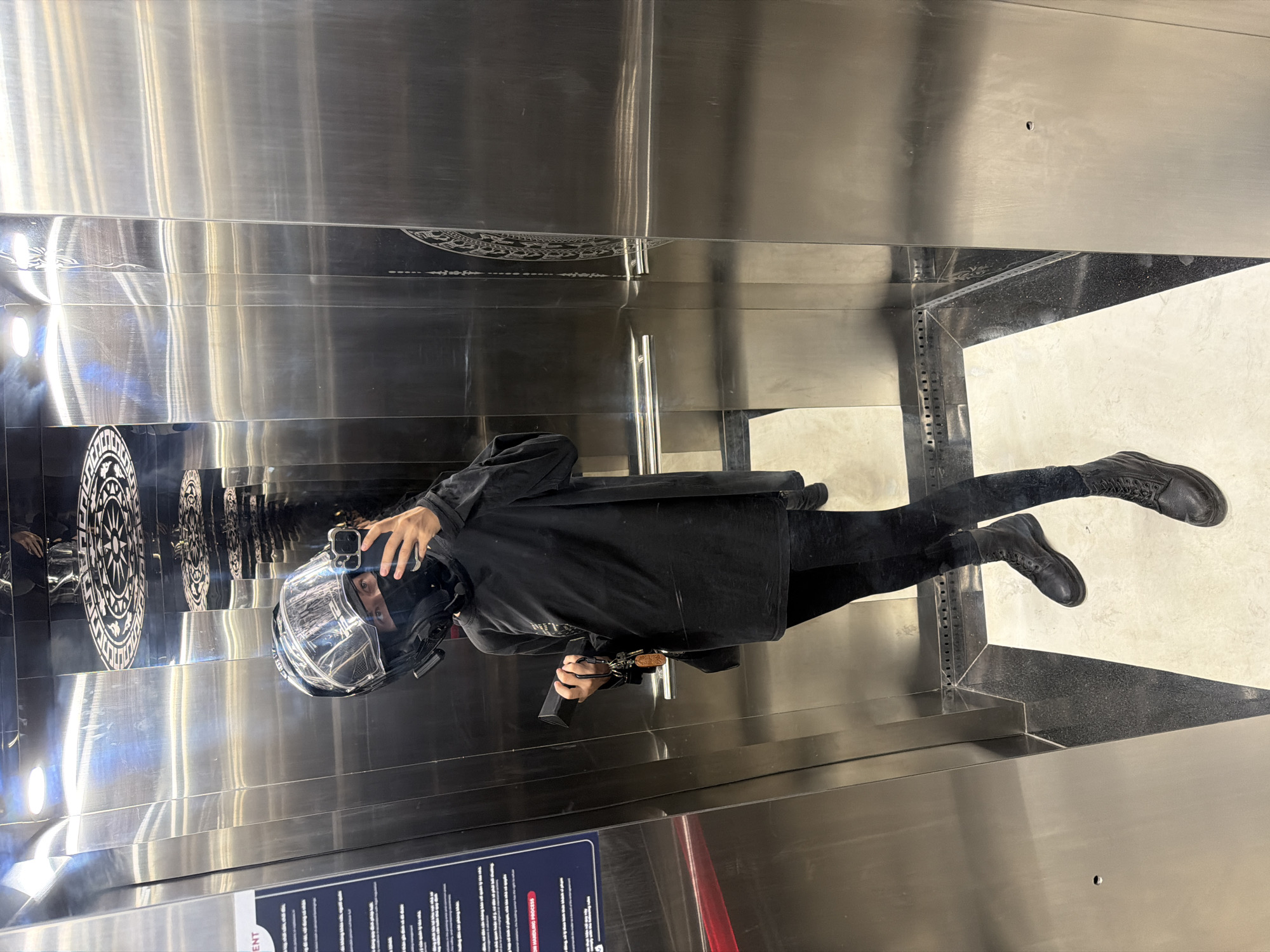 A person wearing a black motorcycle helmet, black jacket, black pants, and black boots is taking a selfie in an elevator. The elevator has stainless steel walls with decorative patterns, including a circular design and intricate lines. A sign with text is visible at the bottom of the elevator wall. The background suggests a clean, modern interior, possibly a shopping mall or commercial building in Đà Nẵng, Vietnam. The person is holding a smartphone in their hand to capture the image.