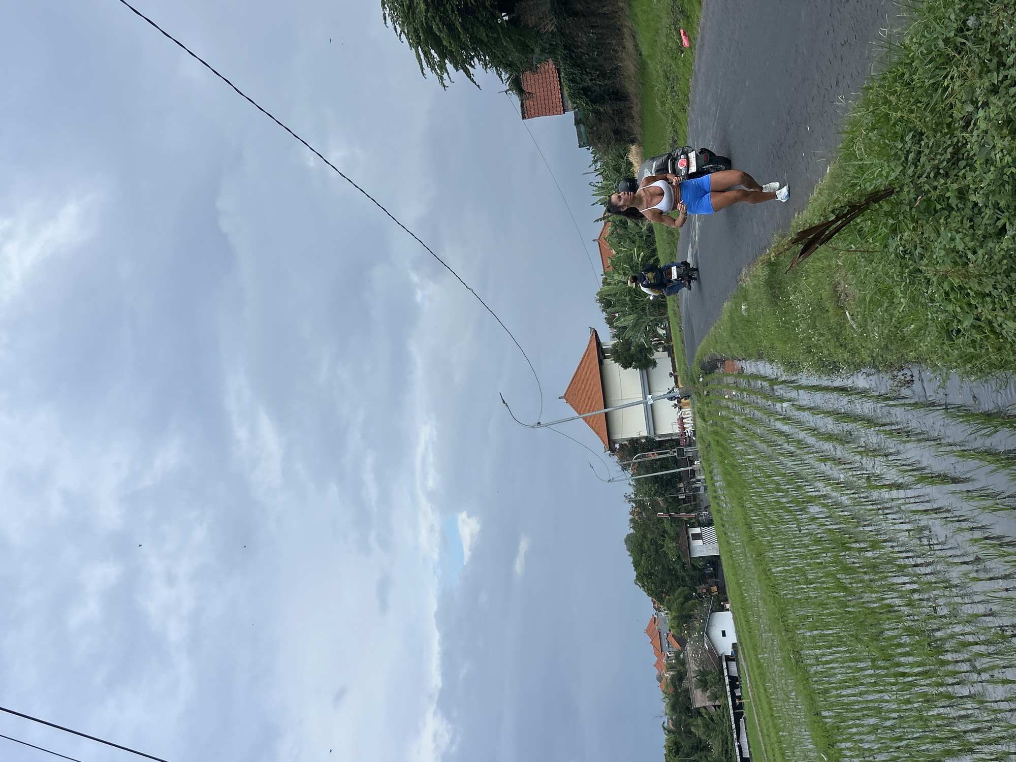 In Mengwi, Indonesia, on an overcast day, a woman runs along a paved road that borders a lush, green rice paddy. She is wearing a white sports bra and blue shorts. Two individuals on motorcycles are visible on the road, one behind the runner and another further ahead. The landscape features a line of buildings with orange-tiled roofs, dense tropical foliage, and a utility pole with wires extending across the sky. The sky is filled with dramatic, gray clouds.
