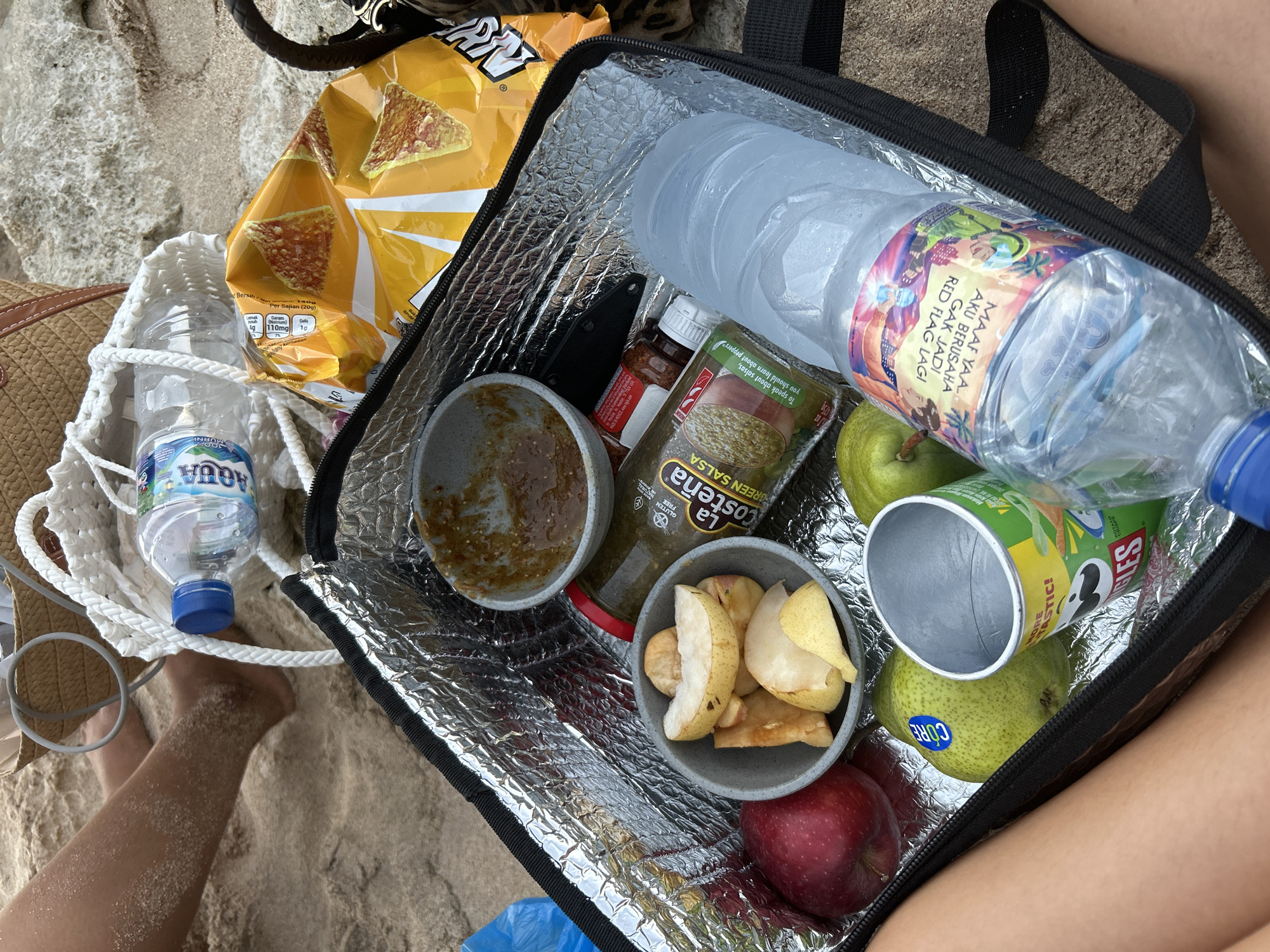 The image depicts a picnic setup on a sandy beach, likely in Kuta Selatan, Indonesia, given the bottle label featuring Indonesian text. The scene is centered around an open, black, insulated cooler bag filled with various food and drink items. Inside the cooler, a grey bowl contains a reddish-brown sauce, possibly salsa, next to a jar of "La Costeña" green salsa. There is also a bowl filled with sliced pears and what appears to be apple slices. A red apple and two green pears are also visible within the bag. A can of Pringles potato crisps is partially visible on the right side. A large plastic bottle of water, condensation indicating it's cold, lies horizontally within the cooler. Outside the bag, a bag of chips, likely Doritos based on the packaging, is placed on the sand. A white woven bag and a partially visible straw bag are also present in the background, along with a blue water bottle with a blue cap. The sand is light brown and appears dry. The lower portion of the image shows the legs of a person, with sand covering their shins and feet. The overall impression is a casual beach picnic with snacks and drinks.