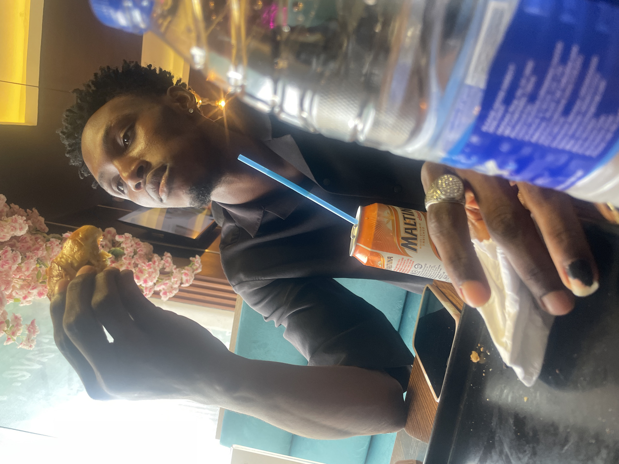 A person is shown from the chest up, eating and drinking at an indoor table. The person has short, curly dark hair and is wearing a dark-colored collared shirt. The person is holding a piece of food in one hand and a napkin in the other. A can of Maltina is on the table with a blue straw inserted. A water bottle is placed near the person. A black phone is lying on the table. The location is Adankolo, Nigeria. Pink floral decorations are visible behind the person.