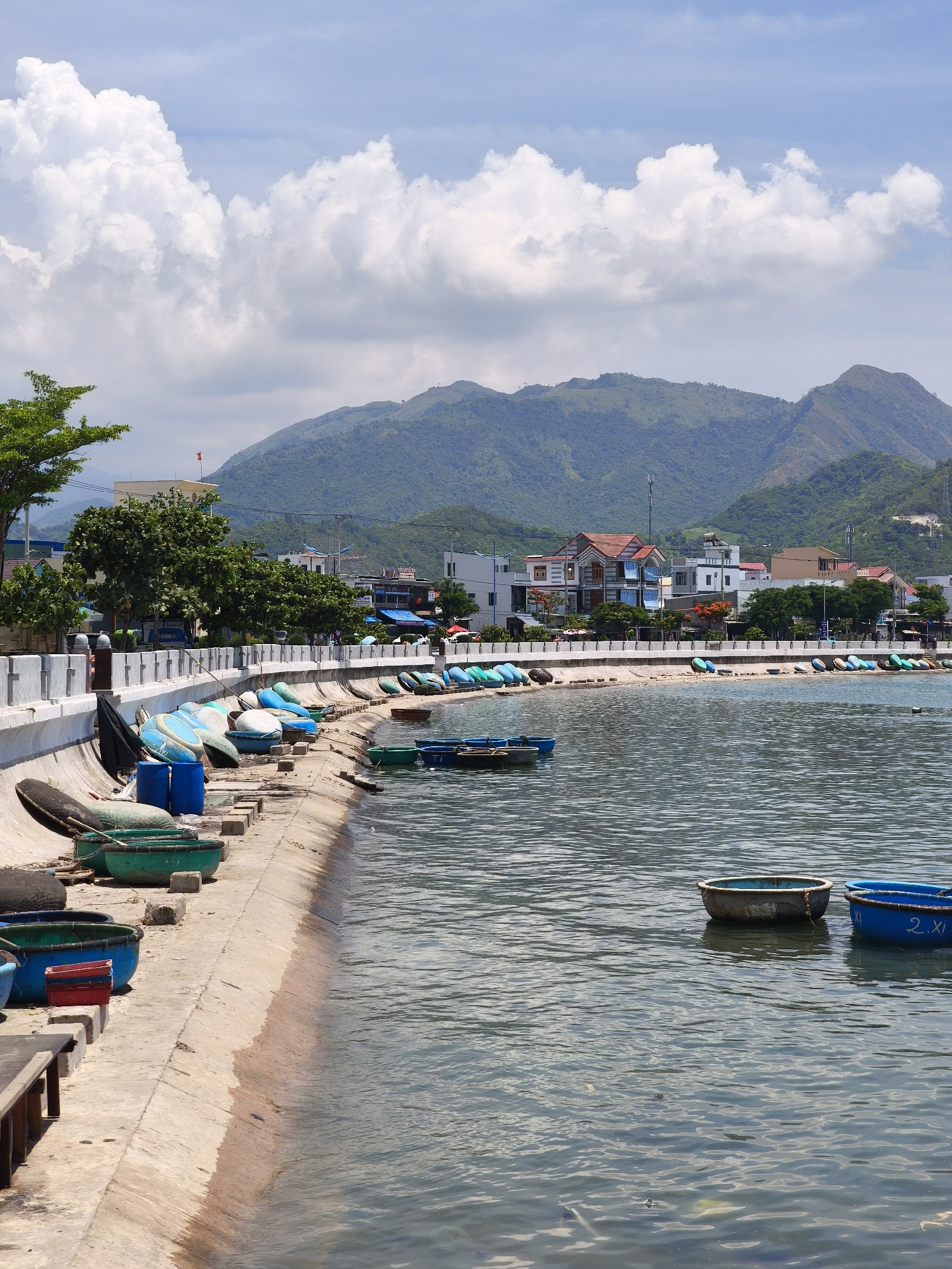 The image captures a coastal scene in Vĩnh Lương, Vietnam, likely on a sunny day given the bright lighting and scattered clouds. In the foreground, the water is calm, with several round, basket-like boats floating and a few more visible on the concrete embankment. These boats are primarily green and blue. The embankment itself is constructed of concrete and stone, showing some wear and tear.
Along the embankment, a collection of blue and white boat hulls are stored, suggesting they are either awaiting repair, storage, or use. Some of these appear to be traditional Vietnamese coracles. There are also a few larger blue barrels and a dark, oval object, possibly another boat or a large piece of equipment, resting on the embankment.
Further back, a paved walkway runs parallel to the shore, separated by a low concrete wall with decorative elements. Beyond the walkway, a line of trees provides a green buffer before the buildings of the town. The town itself consists of low-rise structures, including houses and shops, with various architectural styles. Some buildings feature red tile roofs, while others are modern white or beige structures.
In the background, a range of lush, green mountains rises steeply, forming a natural backdrop to the coastal settlement. The sky is a vibrant blue, punctuated by large, fluffy white clouds. A red flag is visible atop one of the buildings in the town. The overall impression is one of a working coastal community, with evidence of maritime activity and the natural beauty of the Vietnamese coastline.