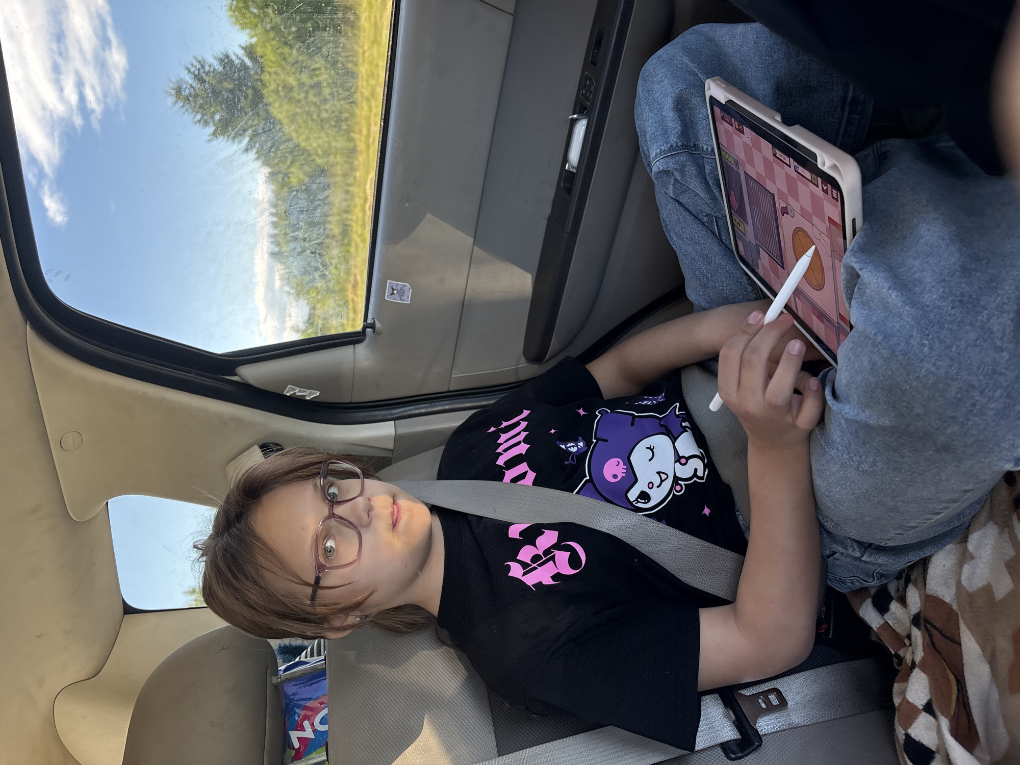 In a car, a young person with brown hair and glasses is lying down, looking at the camera. They are wearing a black t-shirt with a purple cartoon character and pink text. A seatbelt is fastened across their chest. To their right, another person in blue jeans is sitting and using a tablet with a stylus. The tablet screen shows a game with a pink checkered background. Outside the car window, trees and a cloudy sky are visible, suggesting a daytime scene. The interior of the car, with its beige seats and car door panel, indicates the setting is within a vehicle. The overall mood appears relaxed, possibly during a road trip. The scene is likely taking place in Бабаново, Russia.