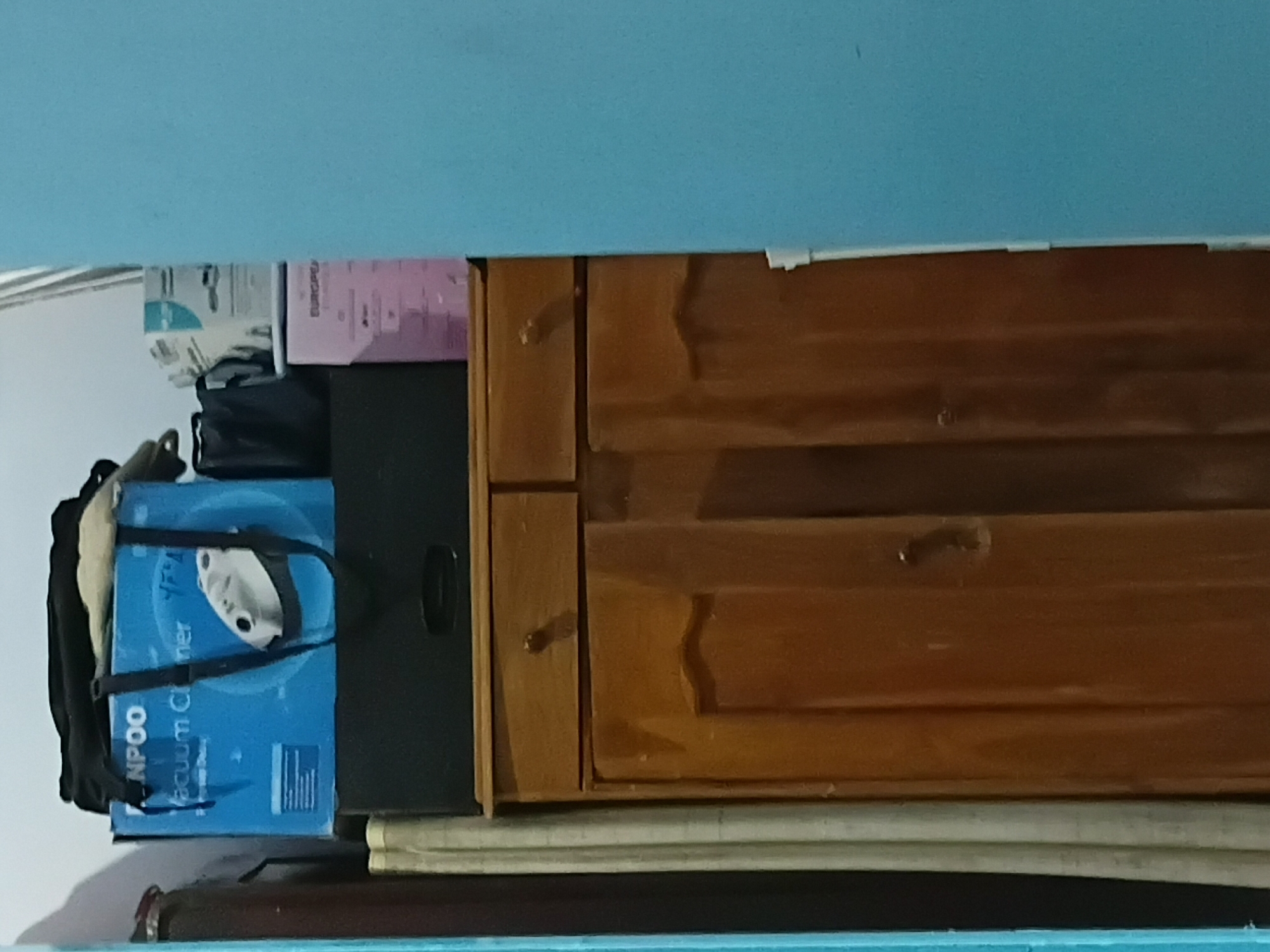 A wooden cabinet with two drawers and two doors stands against a light-colored wall. A dark rectangular object is placed on top of the cabinet. On top of the dark object, there is a blue box with "NPOO Vacuum Cleaner" printed on it. Various other items, including what appear to be bags and boxes, are stacked on top of the blue box. The scene is located in Tanggul Wetan, Indonesia.
