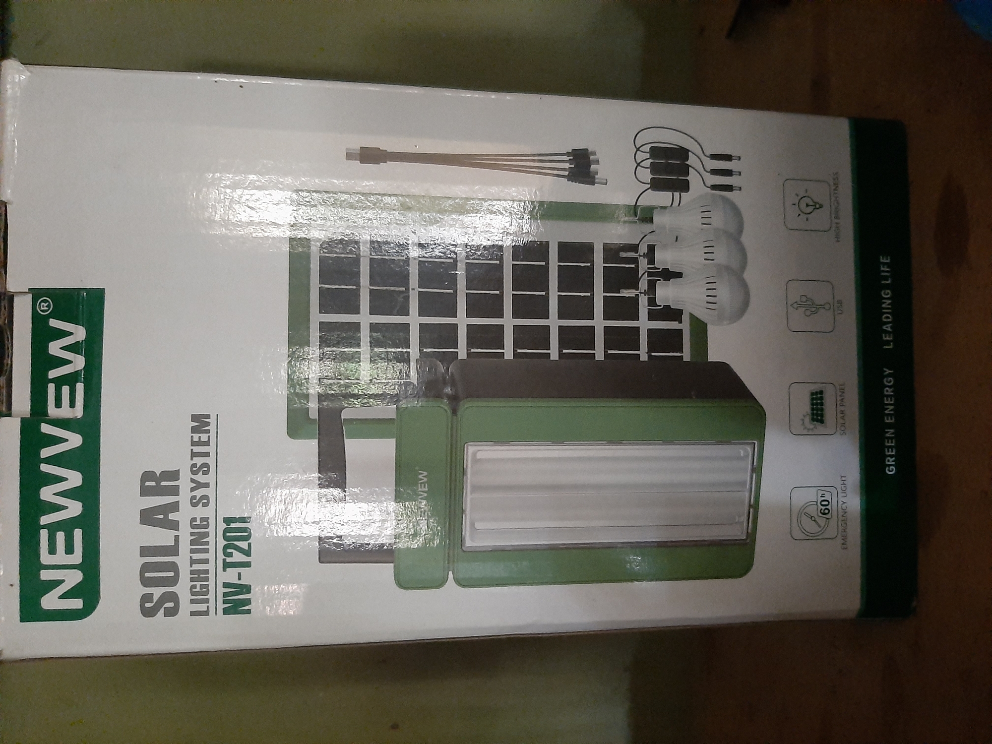 A white cardboard box for a "NEWVEW SOLAR LIGHTING SYSTEM NV-T201" is shown. The box features images of a solar panel, a green lantern-style light with a white LED panel, three hanging light bulbs, and charging cables. Icons indicate "EMERGENCY LIGHT" with "60h", "SOLAR PANEL", "USB", and "HIGH BRIGHTNESS". The bottom of the box reads "GREEN ENERGY LEADING LIFE". The box is positioned on a dark surface, possibly a wooden table, and the background is a mottled green wall. The lighting is somewhat dim, with reflections on the glossy surface of the box. The image appears to have been taken indoors, in a residential or commercial setting. There are no people visible in the image. The scene is in Ogbomosho, Nigeria.