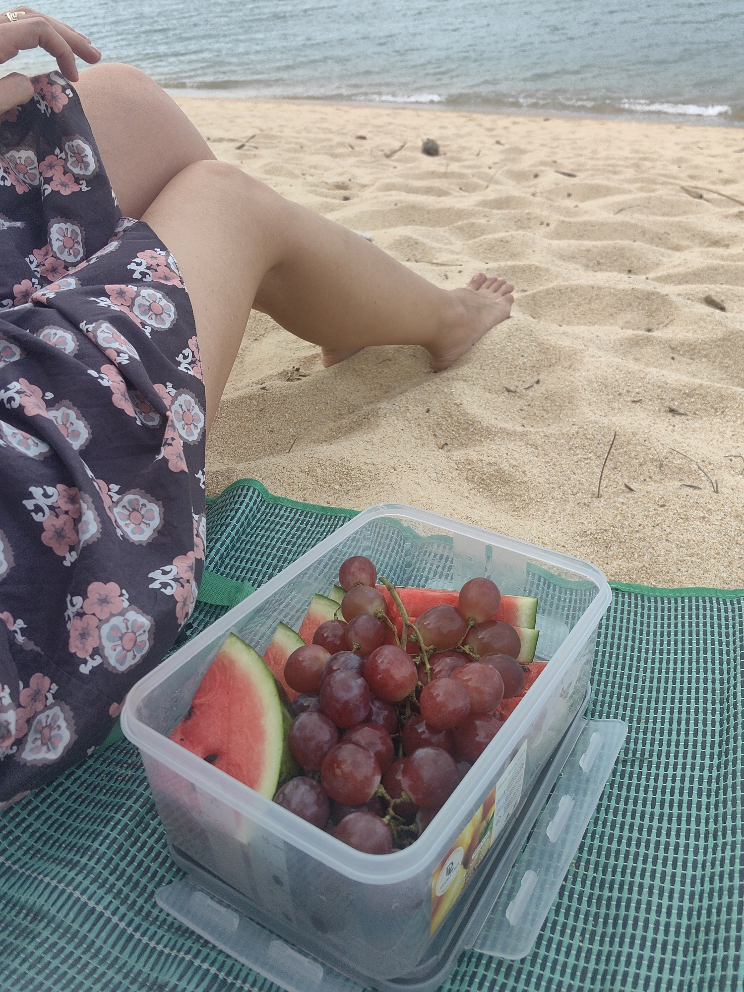 A person sits on a sandy beach in Baan Mae Nam, Thailand, with a picnic container of grapes and watermelon slices to their right. The person's legs, clad in a dark floral skirt, are visible in the foreground, with their feet partially buried in the sand. The ocean stretches out in the background under a hazy sky. The picnic container is made of clear plastic with a white lid. The container rests on a green and black woven mat.