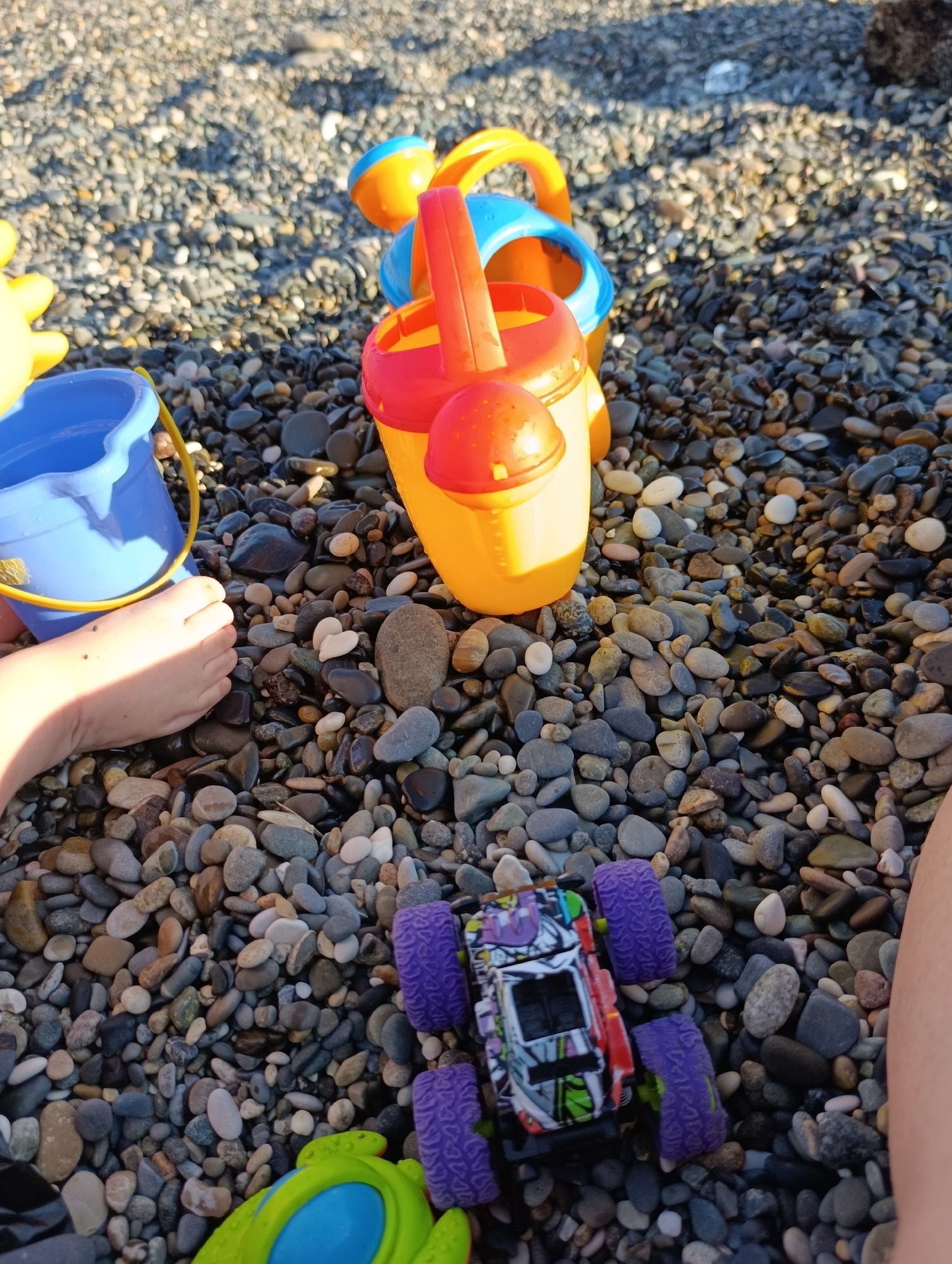 The image shows a close-up of a beach with many small, smooth pebbles. In the foreground, a child's bare foot is visible, with a blue plastic bucket to its left. Behind the foot, several brightly colored plastic beach toys are arranged on the pebbles. These include a yellow and orange watering can with a red handle, and a blue and orange sand mold. Further to the right, a small toy monster truck with purple wheels and a colorful decal is placed on the pebbles. A small portion of another child's leg is visible on the far right of the image. The lighting suggests it is daytime, likely sunny, as there are distinct shadows and bright highlights on the toys and pebbles. The overall scene evokes a sense of play and relaxation, typical of a beach environment. The presence of beach toys and the child's foot strongly indicates that children are playing on the beach. The pebbly shore is a characteristic feature of many coastal areas.