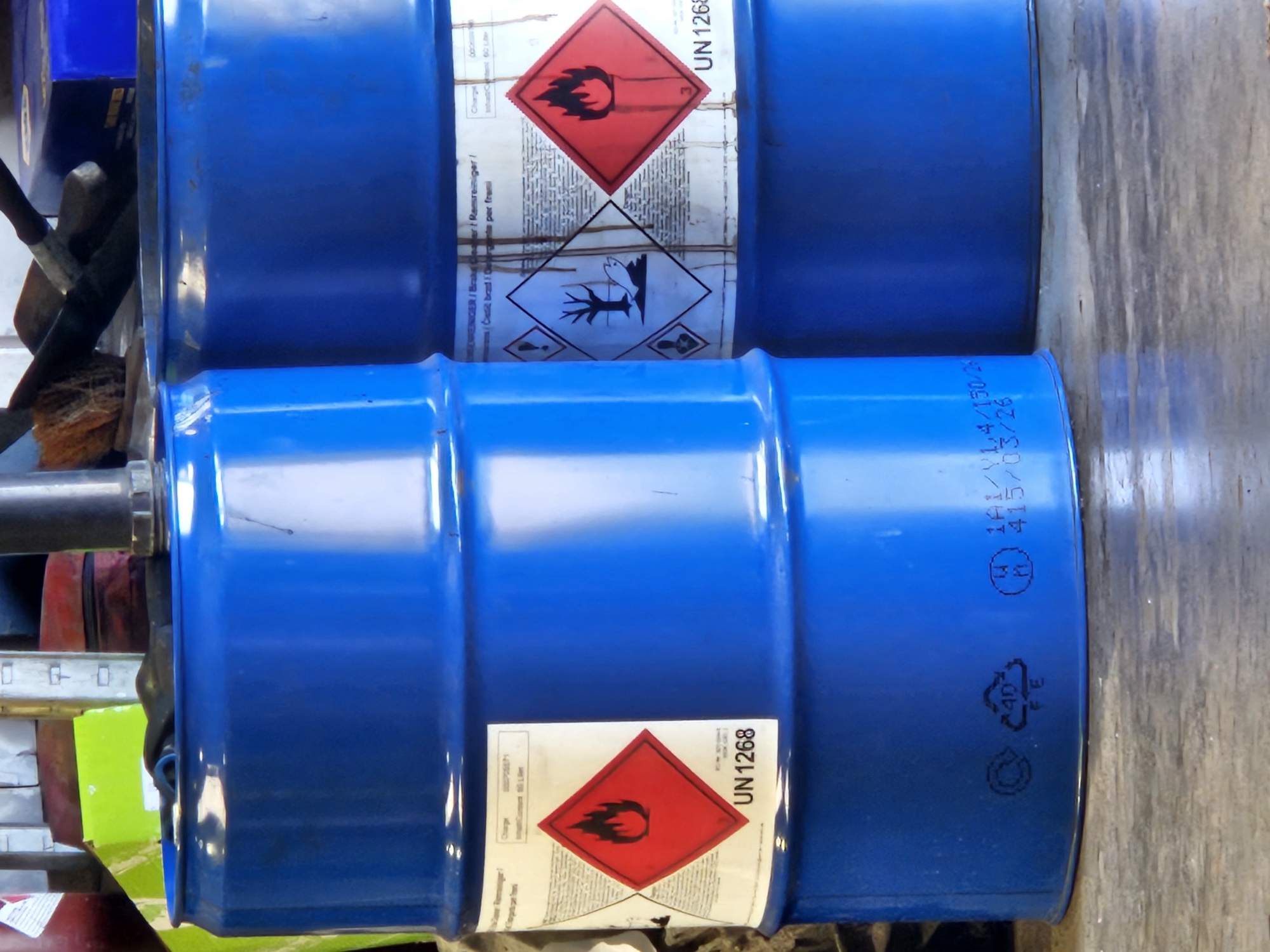 The image shows two blue industrial drums in what appears to be a storage area or workshop. The drum on the left is closer to the viewer and is mostly visible. It has a white label with red hazard symbols and text. Specifically, a red diamond-shaped label with a flame symbol and the text "UN 1268" is prominent. Below this, there are other hazard symbols, including one depicting a dead tree. The drum on the right is partially obscured by the left drum and shows similar labeling. Both drums appear to be made of plastic or metal and are cylindrical. The background is cluttered with various objects, including what looks like tools and other containers, suggesting a functional, possibly industrial or DIY, environment. The lighting is moderate, and the surface beneath the drums appears to be a concrete floor. The image is taken from a slightly low angle, looking up at the drums. The context provided about Bemmel, Netherlands, is external information not directly inferable from the visual content of the image itself.