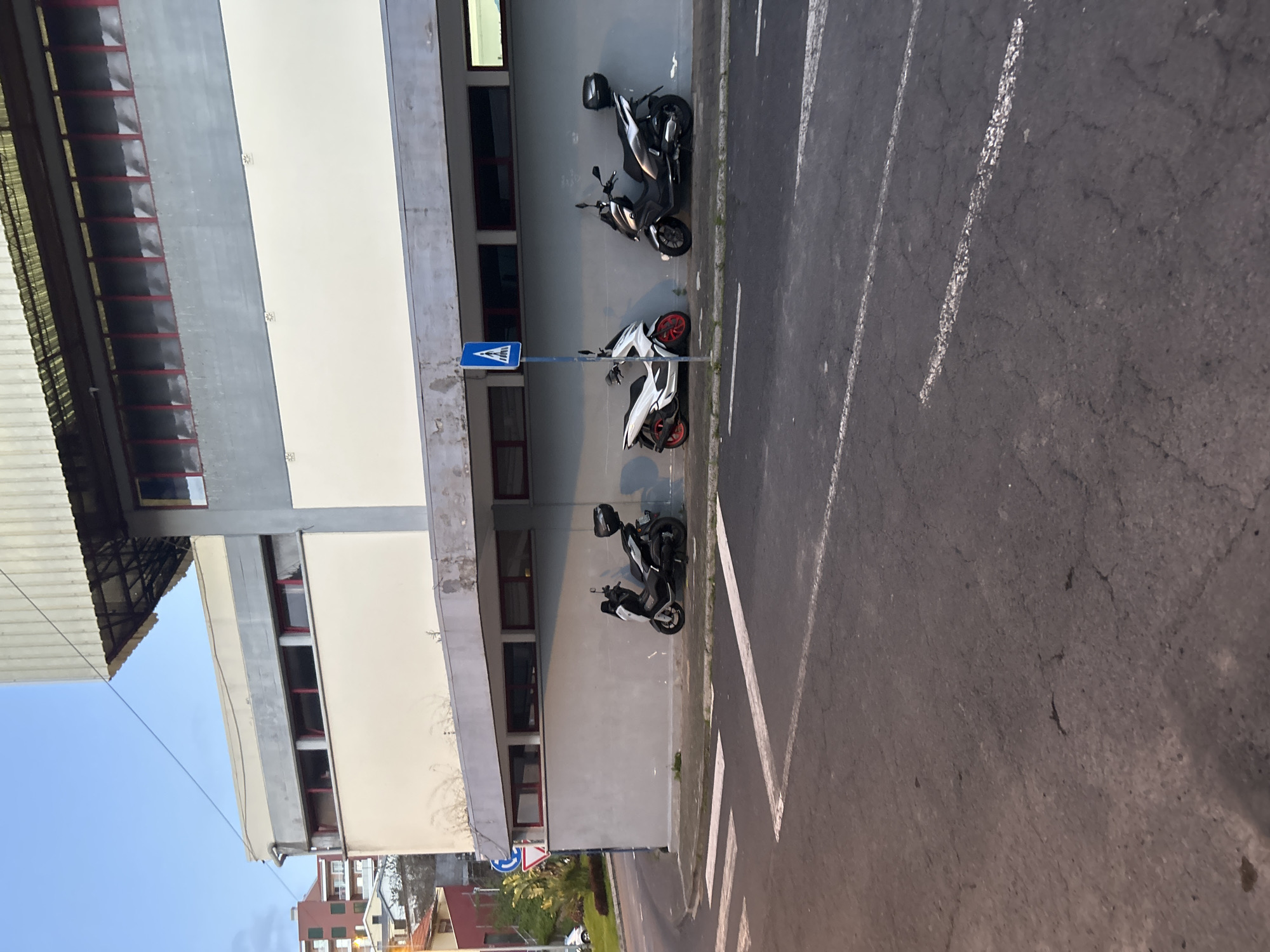 The image shows a street scene in Câmara de Lobos, Portugal. Several scooters are parked against the wall of a multi-story building. The building is light-colored with red trim around the windows. A blue pedestrian crossing sign is visible near one of the scooters. The road is dark gray with white lane markings. The sky is partially visible in the lower-left corner, and a glimpse of other buildings and greenery can be seen in the distance.