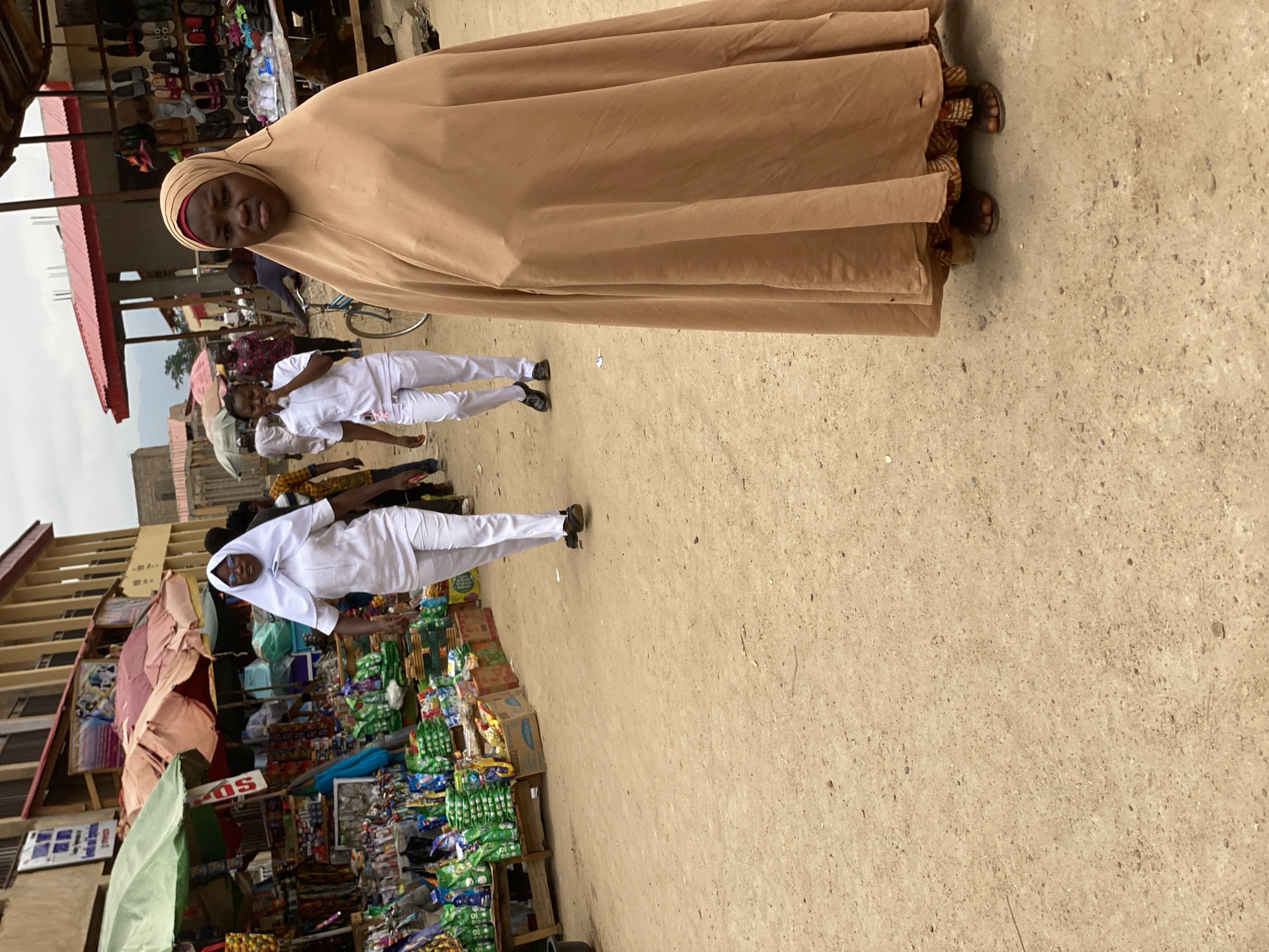 This image captures a vibrant street market scene in Kafanchan B, Nigeria, during daylight hours, likely under an overcast or hazy sky. The ground is unpaved earth.
In the foreground, a woman wearing a long, light brown abaya and hijab stands facing the camera with a neutral expression. She is wearing sandals. Behind her and to the left, two other women are visible, both dressed in white uniforms with white head coverings, suggesting a school or professional uniform. One is walking and looking back, while the other stands near a market stall.
The background is bustling with activity and market stalls. On the far left, a multi-story building can be seen. Various vendors display their goods on the street. One stall features numerous pairs of shoes arranged on shelving. Another stall is densely packed with a wide array of packaged goods, including snacks, drinks, and other small items. A box labeled "Colgate" is visible amongst these products. Several colorful umbrellas provide shade over the merchandise. A red sign with "SO" is partially visible. A bicycle is also present further back in the scene. The overall impression is that of a busy, everyday marketplace.