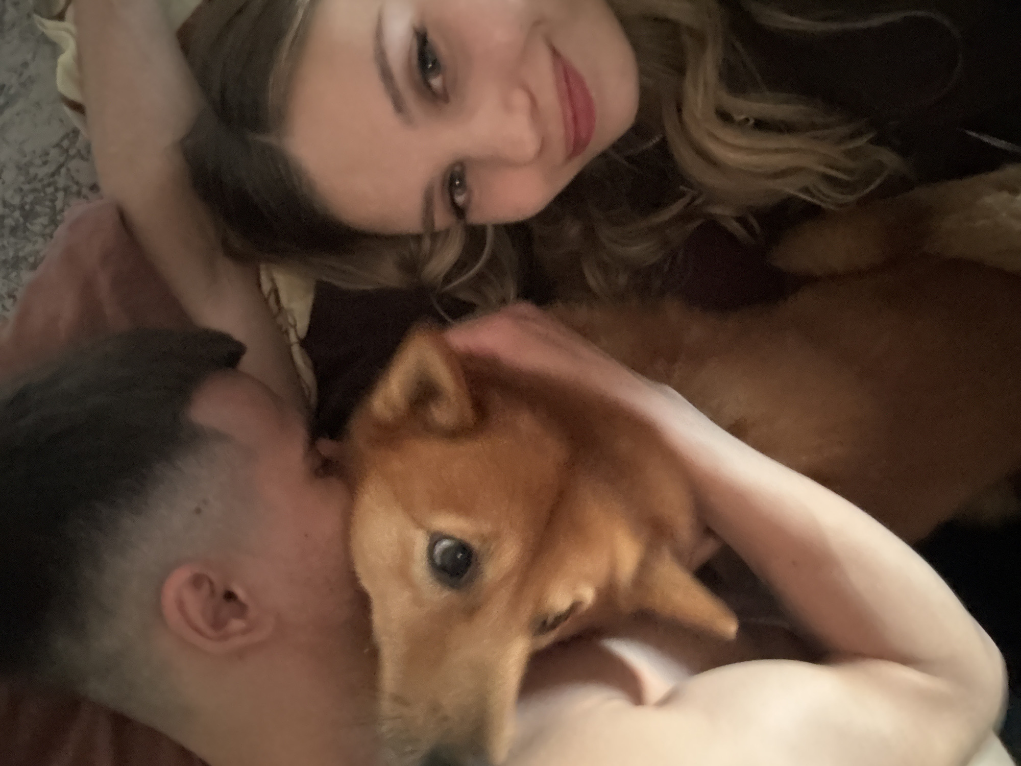 An overhead shot shows a man and a woman lying down, with a dog nestled between them. The man, with a short haircut, is leaning in to kiss the dog. The woman, with blonde hair, is looking at the camera with a smile. The dog, a medium-sized reddish-brown breed, appears to be a Shiba Inu or a similar breed, with large expressive eyes. The group is in a dimly lit room, possibly a bedroom, with a textured grey blanket visible in the background. The image is taken indoors.