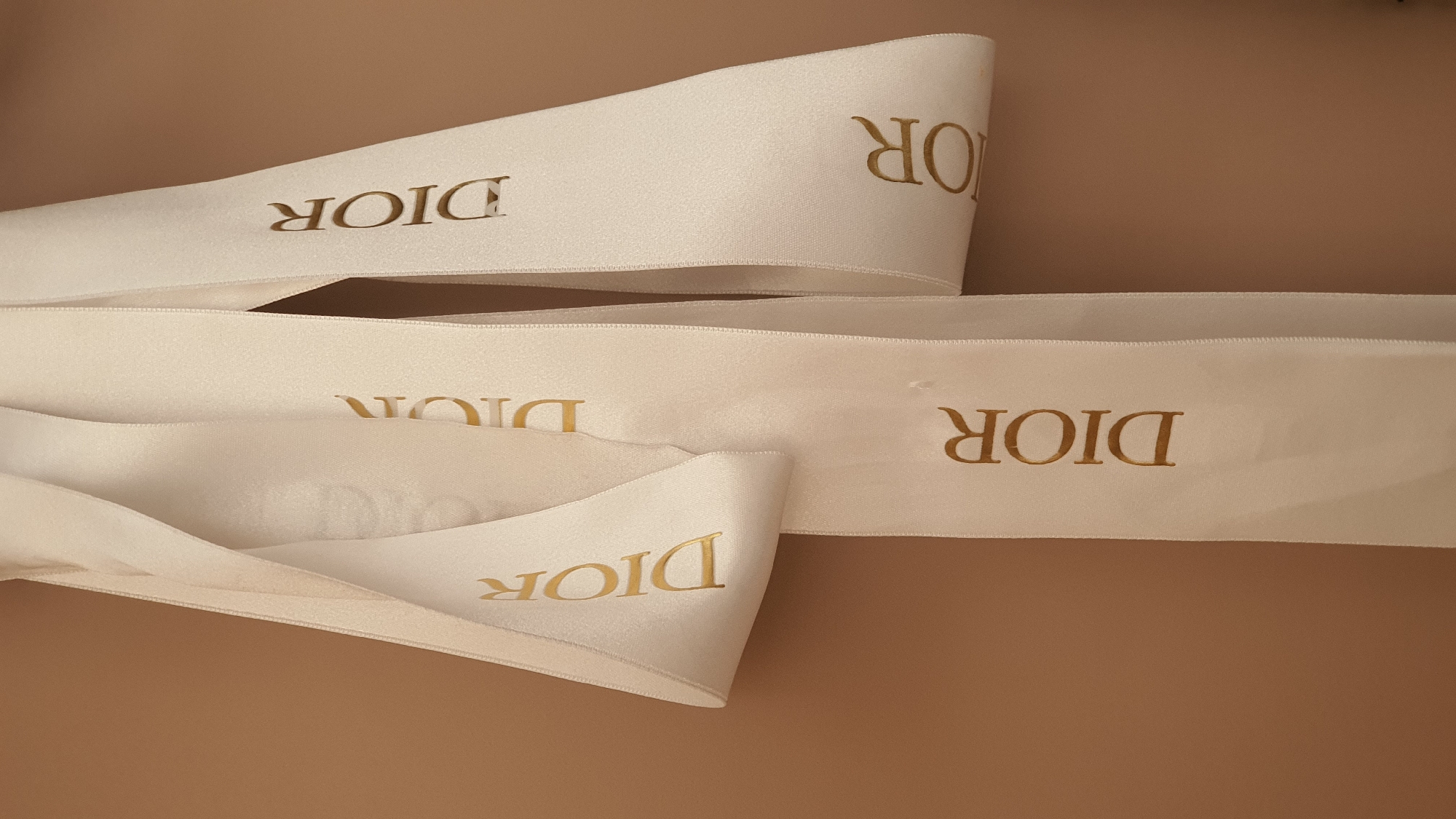 Three white ribbons with the gold lettering "DIOR" are displayed against a brown background. The ribbons are arranged vertically, with the leftmost ribbon folded. The image does not contain any information about the location in the city of Krasnoyarsk, Russia.