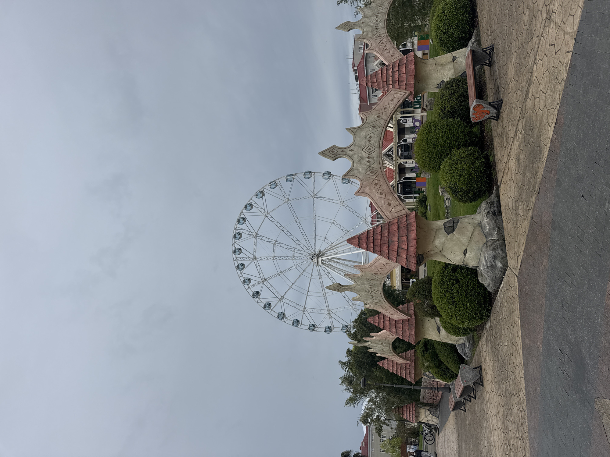 This image, taken in Sochi, Russia, portrays an outdoor amusement or recreational area under an overcast, grey sky during the daytime.
The most prominent subject is a large, white Ferris wheel, which occupies a significant portion of the left side of the frame. Its intricate spoke design and multiple passenger gondolas are clearly visible against the muted sky. To the right of the Ferris wheel, and extending along the right side of the image, are elaborate, themed architectural structures. These feature reddish-brown tiled roofs and light-colored, textured walls adorned with carved geometric patterns, giving them a whimsical or fantasy-like appearance.
A patterned pathway, constructed from interlocking pavers in shades of light brown and darker grey, winds through the foreground and midground on the right. Along this path and nestled among the decorative structures are numerous well-maintained, rounded green bushes and some trees. Several park benches, made of wooden slats with dark metal frames, are placed along the pathway; one notable bench has an orange, abstract design on its side panel.
In the background, behind the immediate attractions, multi-story buildings, some with red roofs, can be seen, suggesting a surrounding urban or resort environment. A few indistinct figures of people are barely visible in the far distance near these buildings, and a bicycle is seen near a small person on the main path, indicating some pedestrian activity. However, no specific event or intense activity is taking place in the foreground. No legible text is visible on any signs or objects within the frame. The overall atmosphere is serene and calm due to the cloudy weather and the absence of bustling crowds in the foreground.