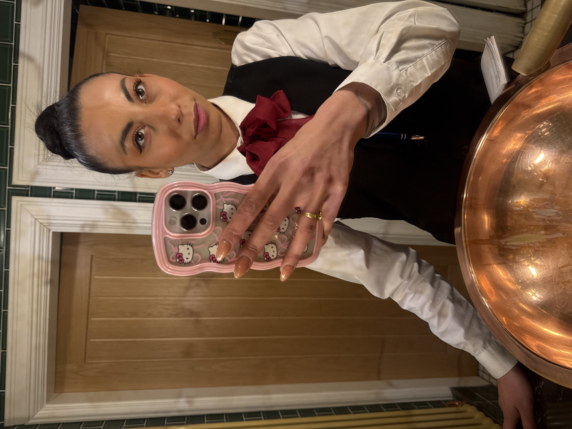 The image is a close-up selfie of a woman wearing a white button-down shirt with a black vest and a dark red bow tie. Her dark hair is pulled back in a bun. She is holding a pink smartphone case with Hello Kitty designs. A large copper bowl with reflective interior is visible on the right. In the background, there is a door and wall paneling with green tile accents. There is no definitive information to establish the location in Warrington, United Kingdom.