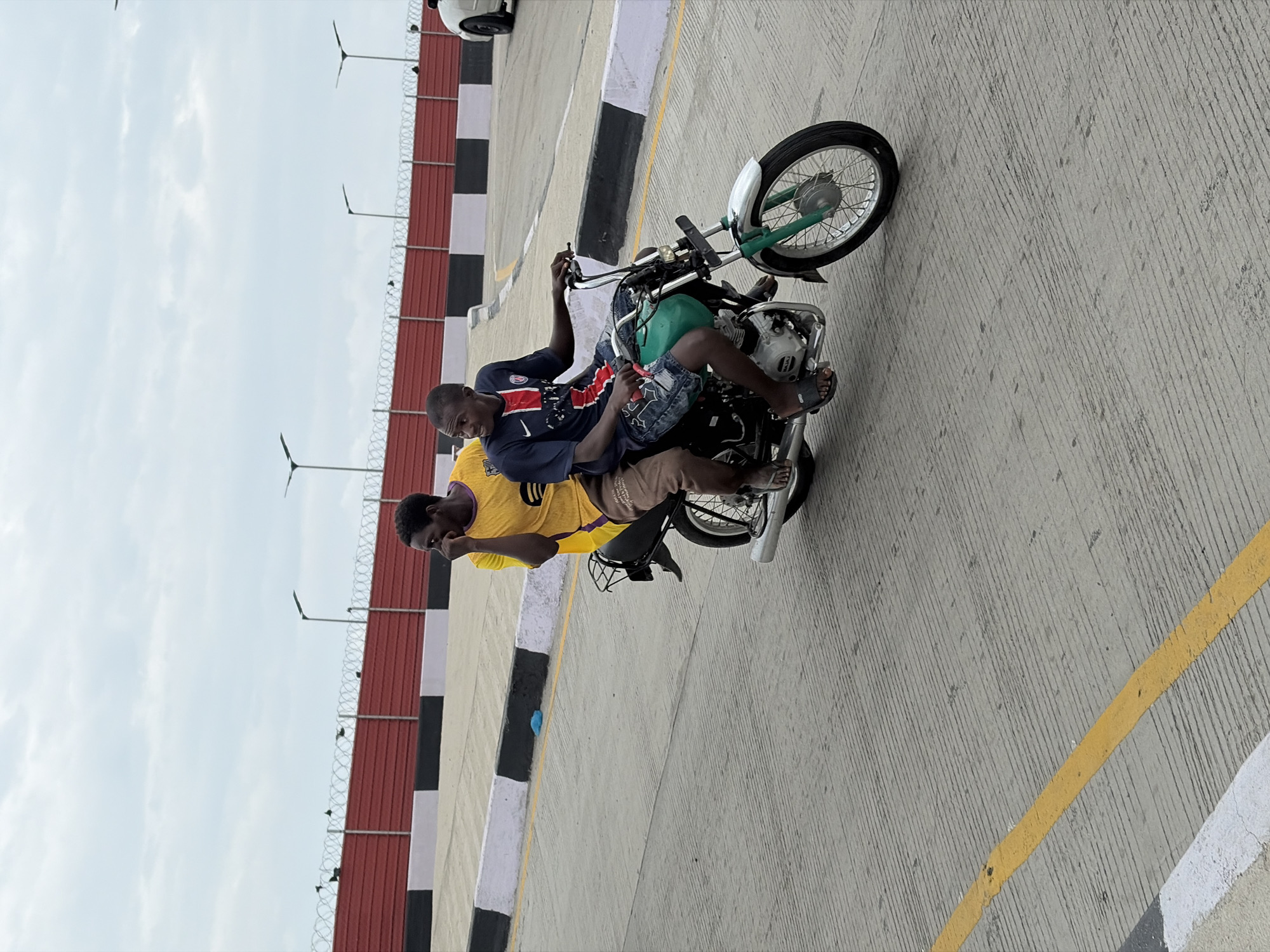 Two men are riding a motorcycle on a paved road in Alaguntan, Nigeria. The man in the front wears a yellow shirt and khaki pants. The man behind him wears a blue shirt and patterned denim shorts. Both men are wearing sandals. The motorcycle is dark green and silver. The road has yellow and white lines. A fence with razor wire is visible along the road. Street lights are also visible.