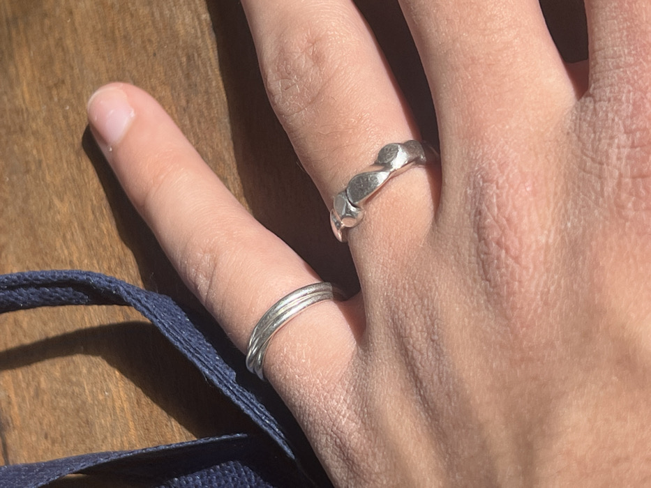 The image displays a close-up, high-angle view of a person's left hand resting on a wooden surface, located in the city of Hof Ashkelon Regional Council, Israel. The hand's skin tone is light, with visible skin texture and creases. Two silver-toned rings are worn on the hand: one on the ring finger, consisting of three thin, parallel bands that appear twisted or stacked; and another on the middle finger, featuring an undulating, patterned design with rounded, raised elements. Strong, natural light illuminates the scene, casting distinct shadows of the fingers and rings onto the wooden surface. A dark blue, woven fabric strap or cord is visible in the upper left portion of the frame, partially out of focus, lying on the wooden surface. The wooden surface itself exhibits a visible grain pattern.