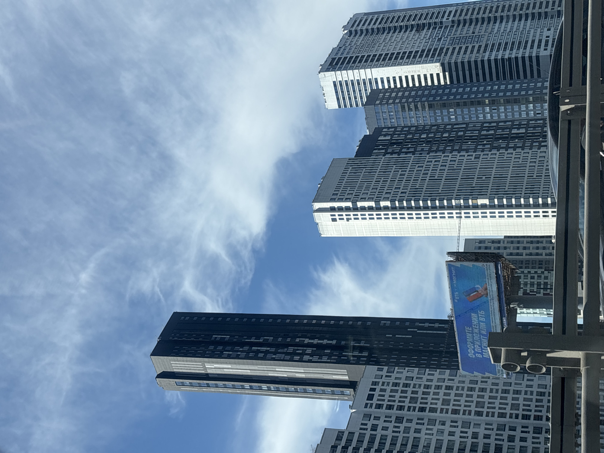 The image shows a view of several modern high-rise buildings against a blue sky with wispy clouds. In the foreground, there is a metal barrier. A billboard is visible in the middle ground, displaying an advertisement with Russian text and images of mobile phones. The buildings are a mix of dark and light facades, with numerous windows. The overall scene depicts an urban environment, likely a business or residential district, in Moscow, Russia.