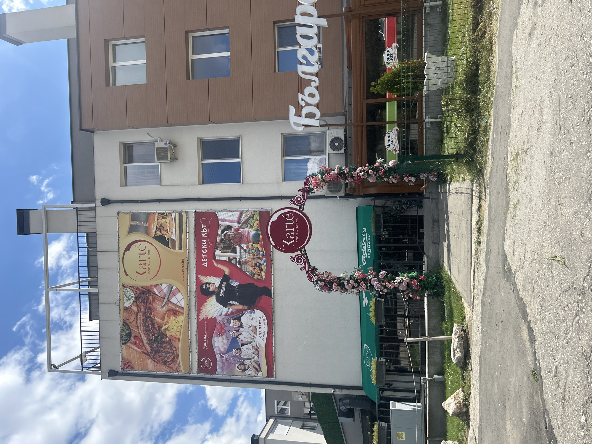 The image captures the exterior of a building in Varna, Bulgaria, likely a restaurant or entertainment venue named "Karte Food & Drink". The building has multiple levels, with large advertising banners displayed on the facade. One banner showcases a large steak and a burger, while another features children in a ball pit and a woman with wings, promoting "Spa Party for Little Princesses" and "Kids Corner". A floral archway decorated with pink and white flowers leads to the entrance of the venue, which has a green awning. The name "Болгаро" (Bulgaro) is visible on a prominent sign. In front of the building, there is a paved walkway, grass, and some rocks. The overall scene suggests a commercial establishment with offerings for both adults and children.