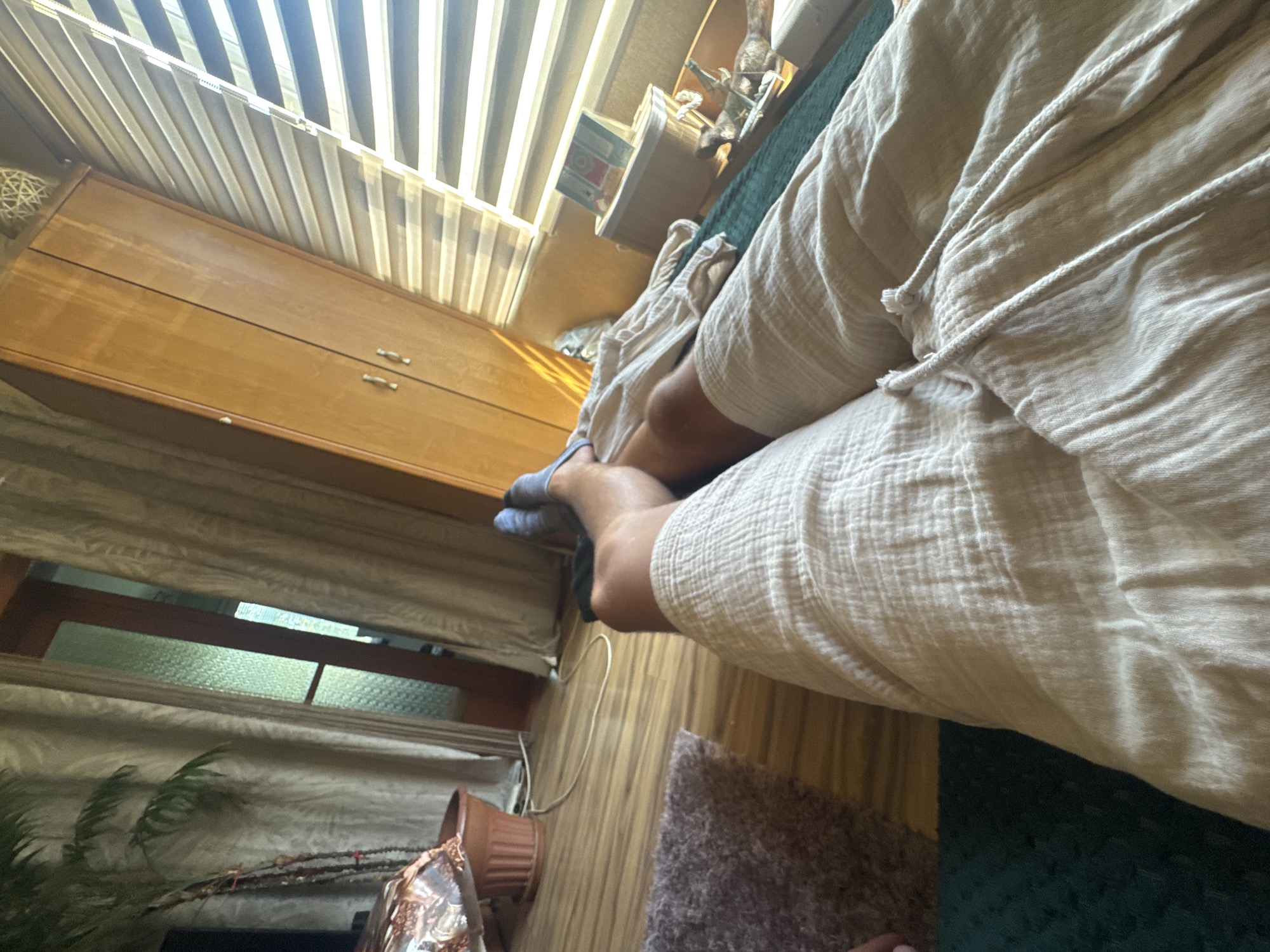 The image shows a first-person perspective of a person lying down, with their legs covered by light-colored, loose-fitting shorts and blue socks. The person is lying on a dark green surface, possibly a bed or sofa, with a glimpse of a patterned rug and wooden flooring. In the background, a wooden wardrobe with two doors and silver handles is visible. To the right of the wardrobe, a window with horizontal blinds is letting in sunlight, casting striped shadows. A tall houseplant in a terracotta pot stands to the left, near a door with frosted glass panels and beige curtains. The scene appears to be an indoor setting, likely a bedroom or living room. The image does not contain enough information to determine its location in Sofia, Bulgaria.