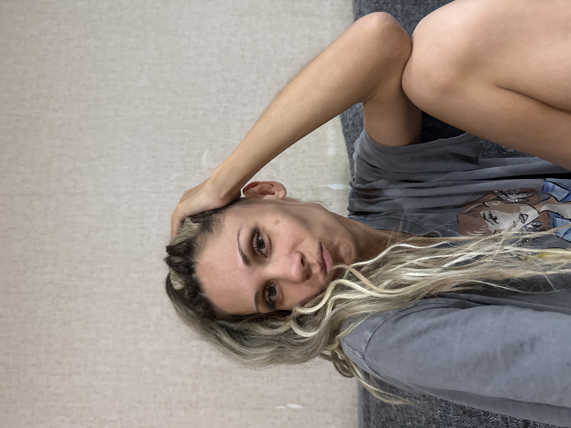 A medium shot shows a woman with blonde dreadlocks and dark roots, looking at the camera with her right hand resting on her head. She is wearing a gray t-shirt with a graphic print of a woman with purple earrings and blonde hair. The woman's lips are pursed, and her expression is somewhat bored or contemplative. She is sitting on a gray couch against a textured beige wall. The lighting is soft and even. No specific location in Rusanovo, Russia is evident from the image.