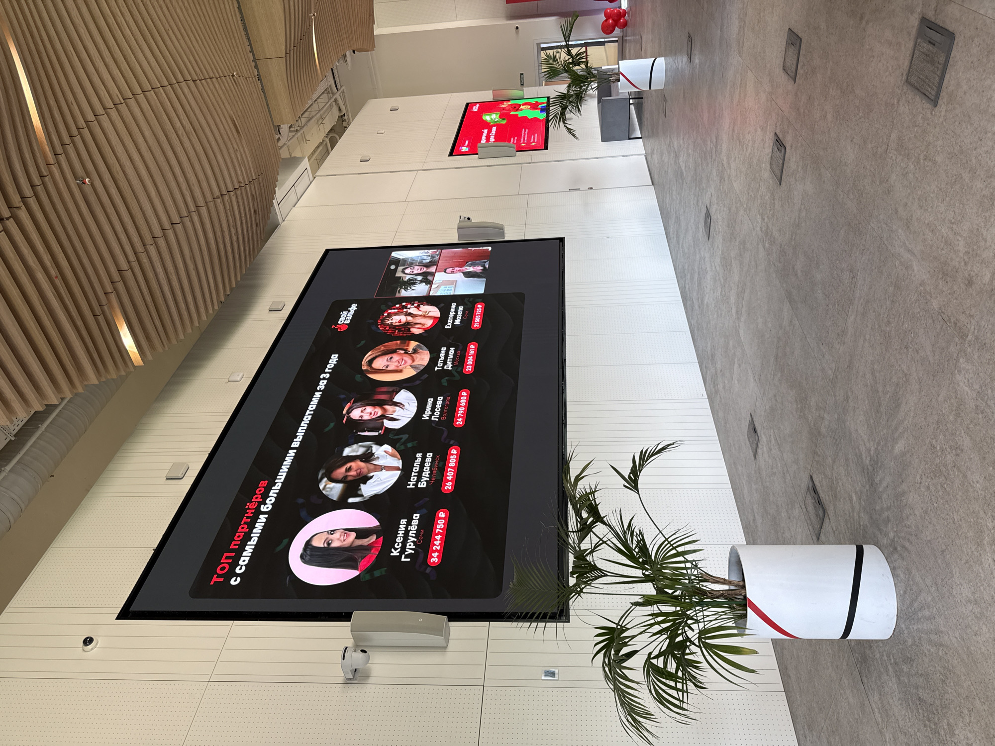 Here is a description of the image:
The image depicts the interior of a modern building, possibly an office or commercial space. A large digital screen is mounted on a white wall, displaying what appears to be a ranking of top partners with the highest payouts over three years, featuring photos and names of people, along with monetary amounts in rubles (P). The text on the screen is in Russian. Above this screen, there's another smaller screen displaying content, possibly promotional.
To the right of the main screen, a plant in a cylindrical white pot with colored stripes adds a touch of greenery to the space. The flooring is a light-colored tile. The lighting appears to be bright, and the architecture features modern, clean lines.
Based on the names listed on the screen, "Sochi" and "Moscow" are listed as locations for some of the people featured on the screen, indicating the location could be Russia, potentially in Moscow, but not confirmed with certainty based on the image.